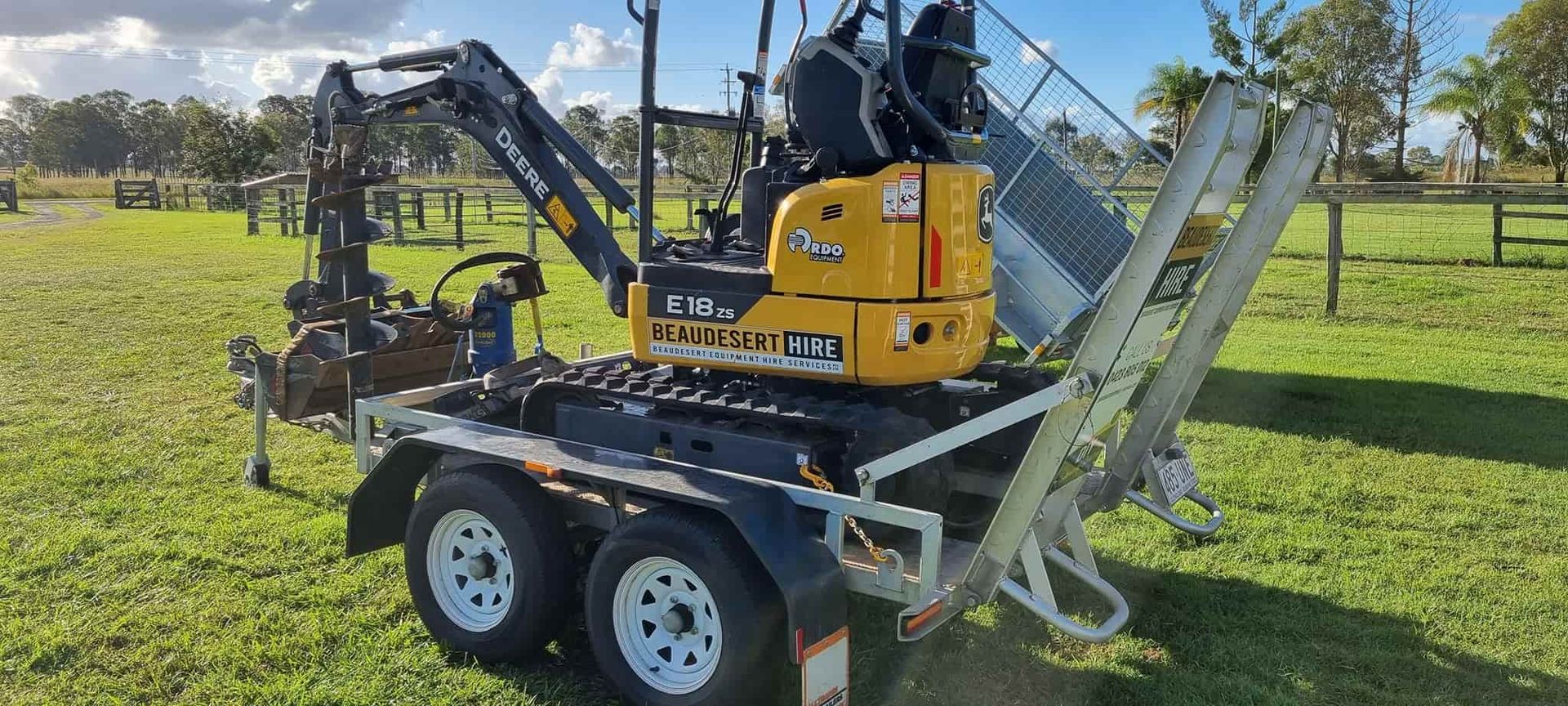 A small yellow excavator is parked on a trailer in a grassy field. — Beaudesert Equipment Hire Services in Beaudesert, QLD