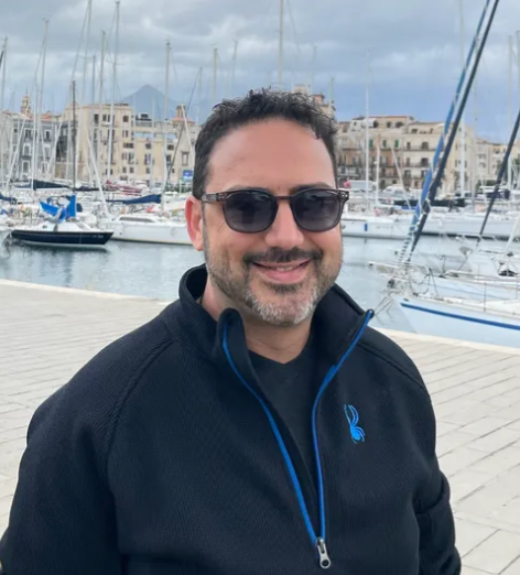A man wearing sunglasses and a black jacket stands in front of boats