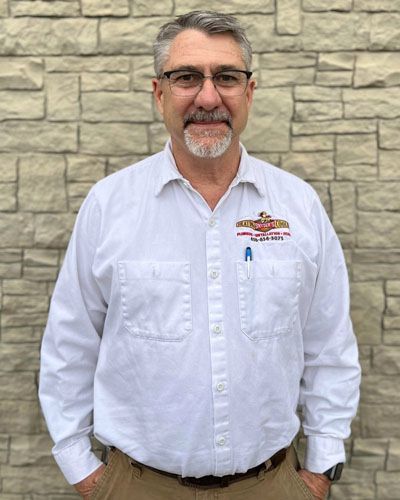 A man wearing glasses and a white shirt is standing in front of a brick wall.