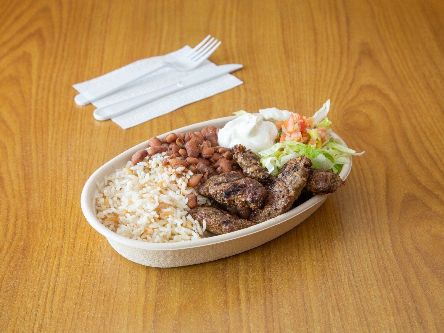 A bowl of food with rice , meat , beans and salad on a wooden table.