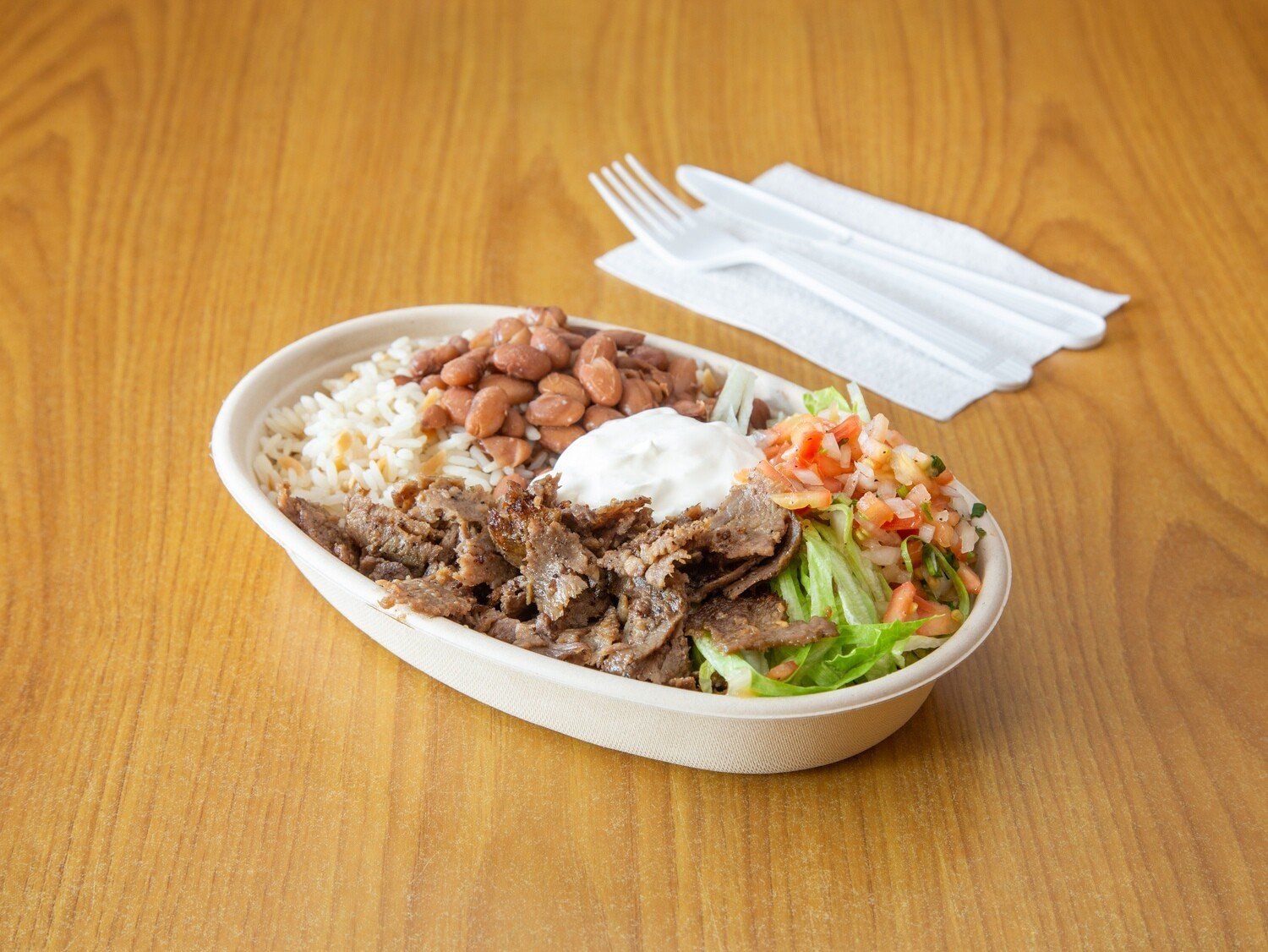 A bowl of food with rice , beans , tomatoes and sour cream on a wooden table.