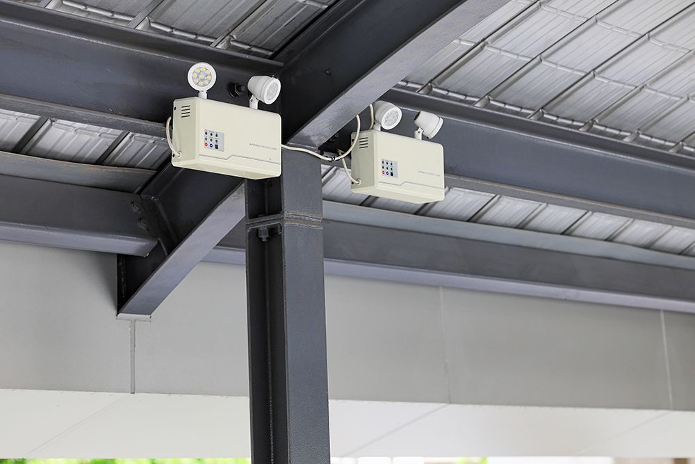 Two beige emergency lights mounted on a metal beam, under a corrugated metal roof.