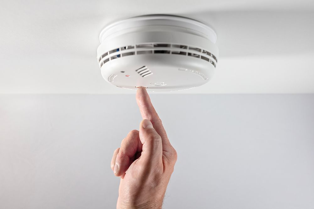 A person testing a white smoke detector on a ceiling with their finger.