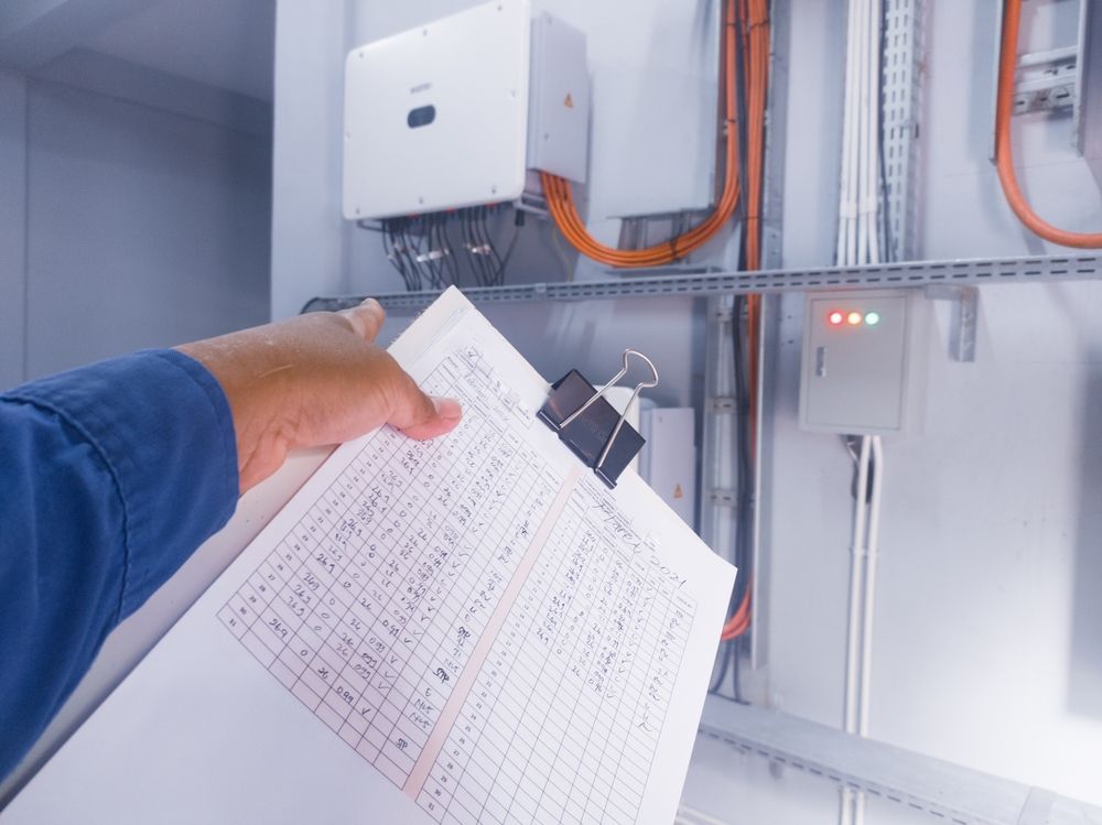 A person holding a clipboard inspecting electrical equipment with wires and lights.