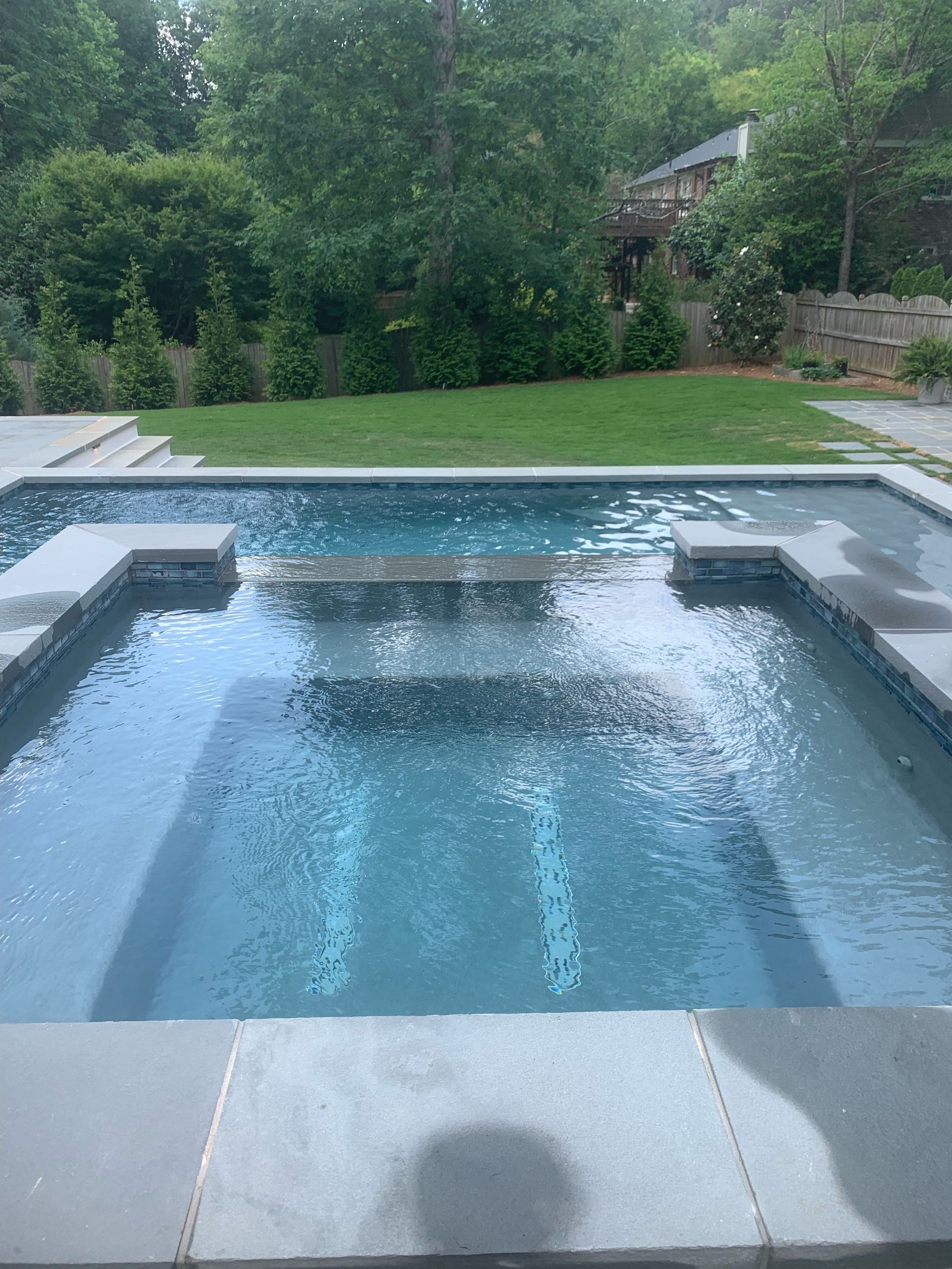 A backyard swimming pool with an attached spa, framed by gray stone coping, overlooking a green lawn and trees.