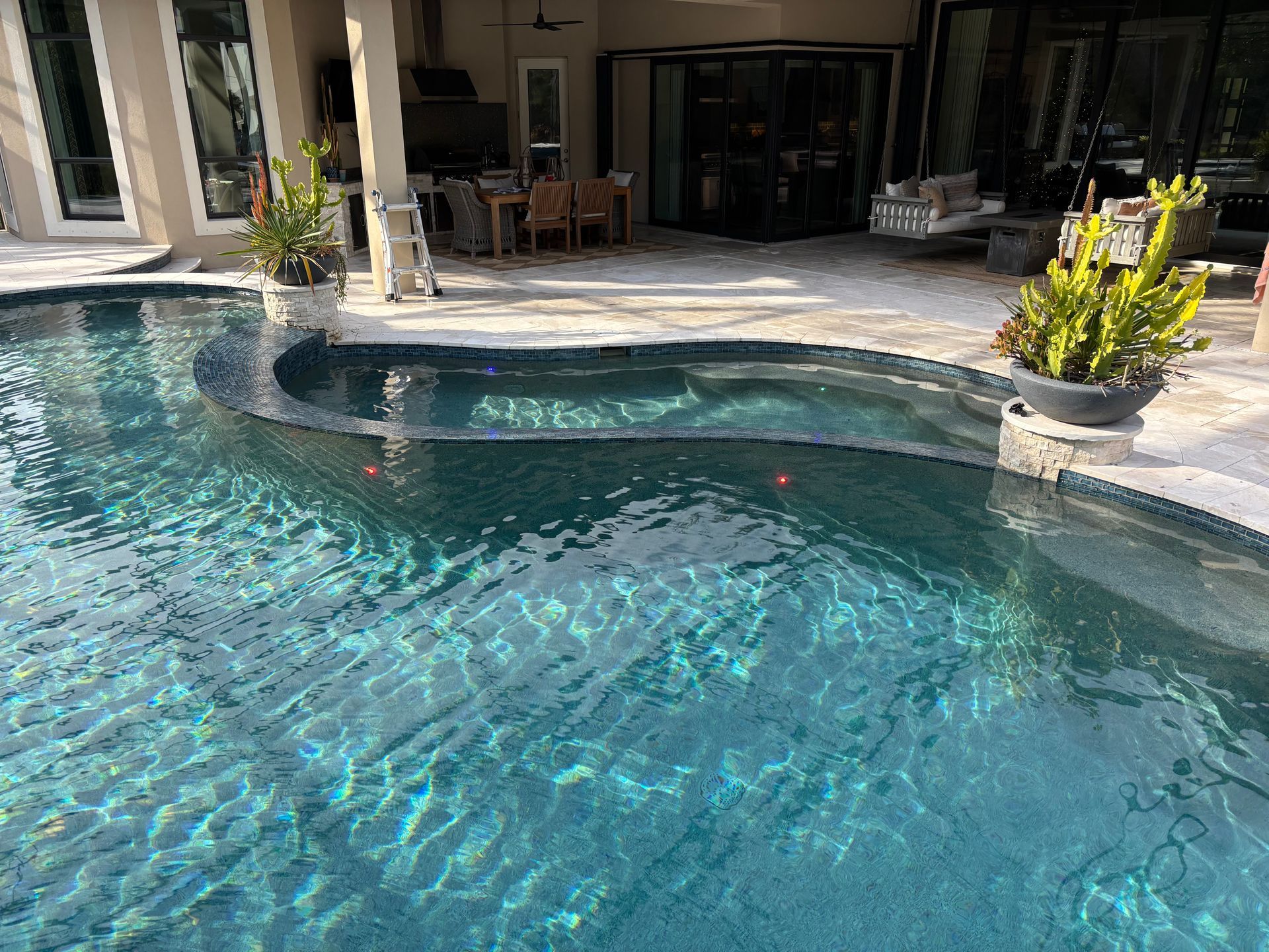 A clear swimming pool with a curved raised spa edge, overlooking a patio area with outdoor furniture and a house.