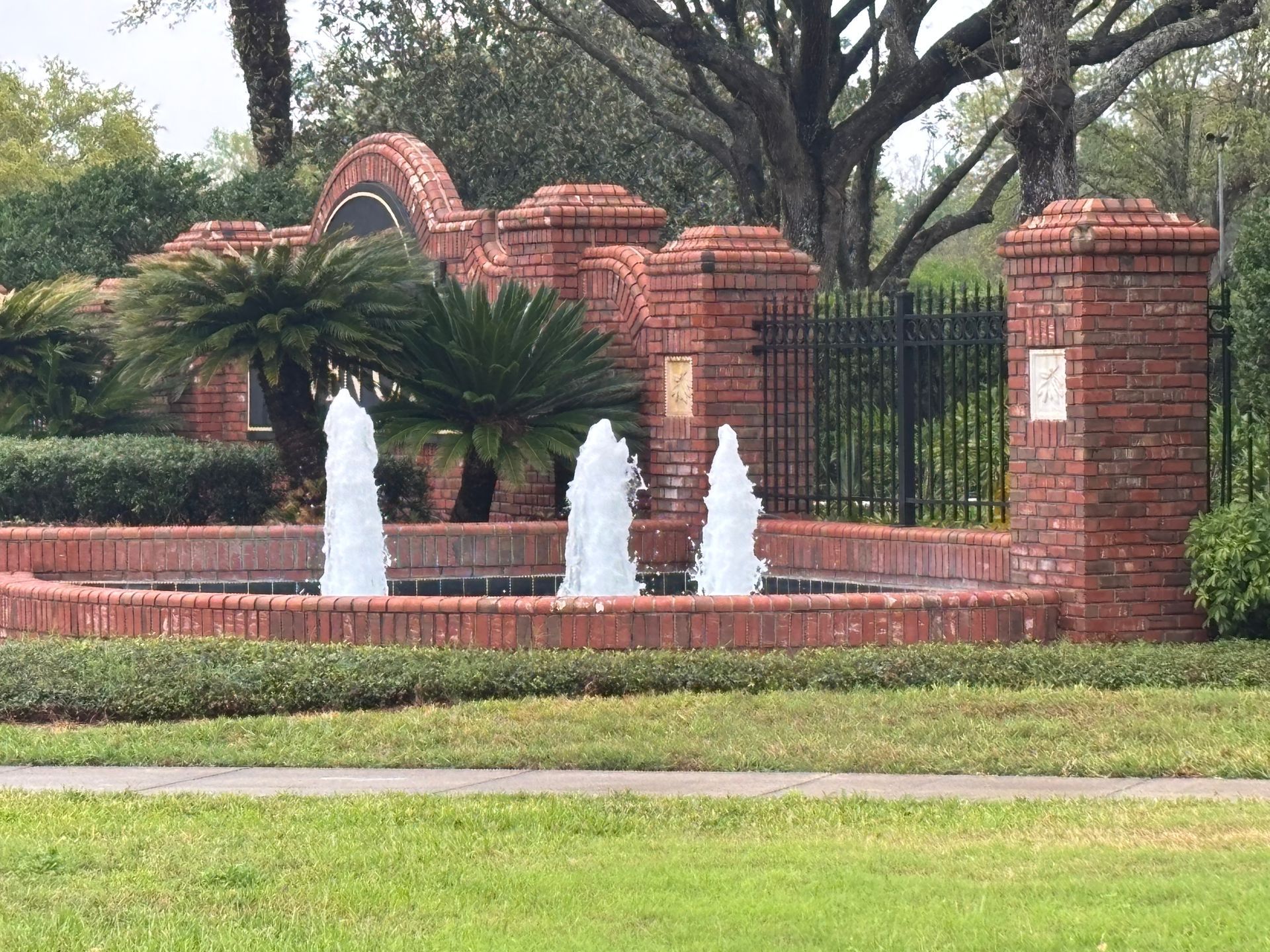 A brick neighborhood entrance with a circular fountain spraying three jets of water in front of palm trees and black fencing.