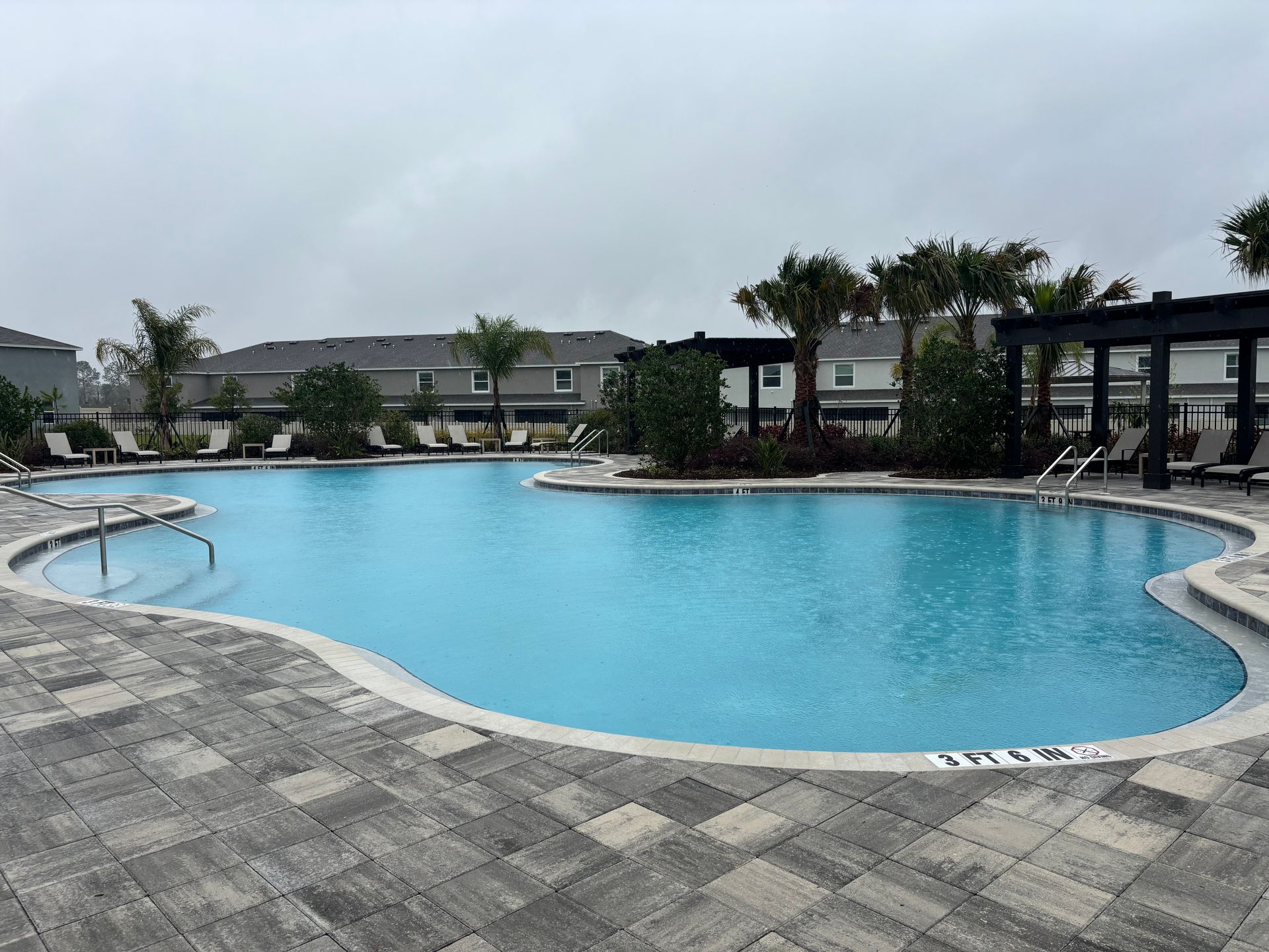 A curved outdoor swimming pool with blue water and stone pavers, surrounded by palm trees and pergolas under a cloudy sky.