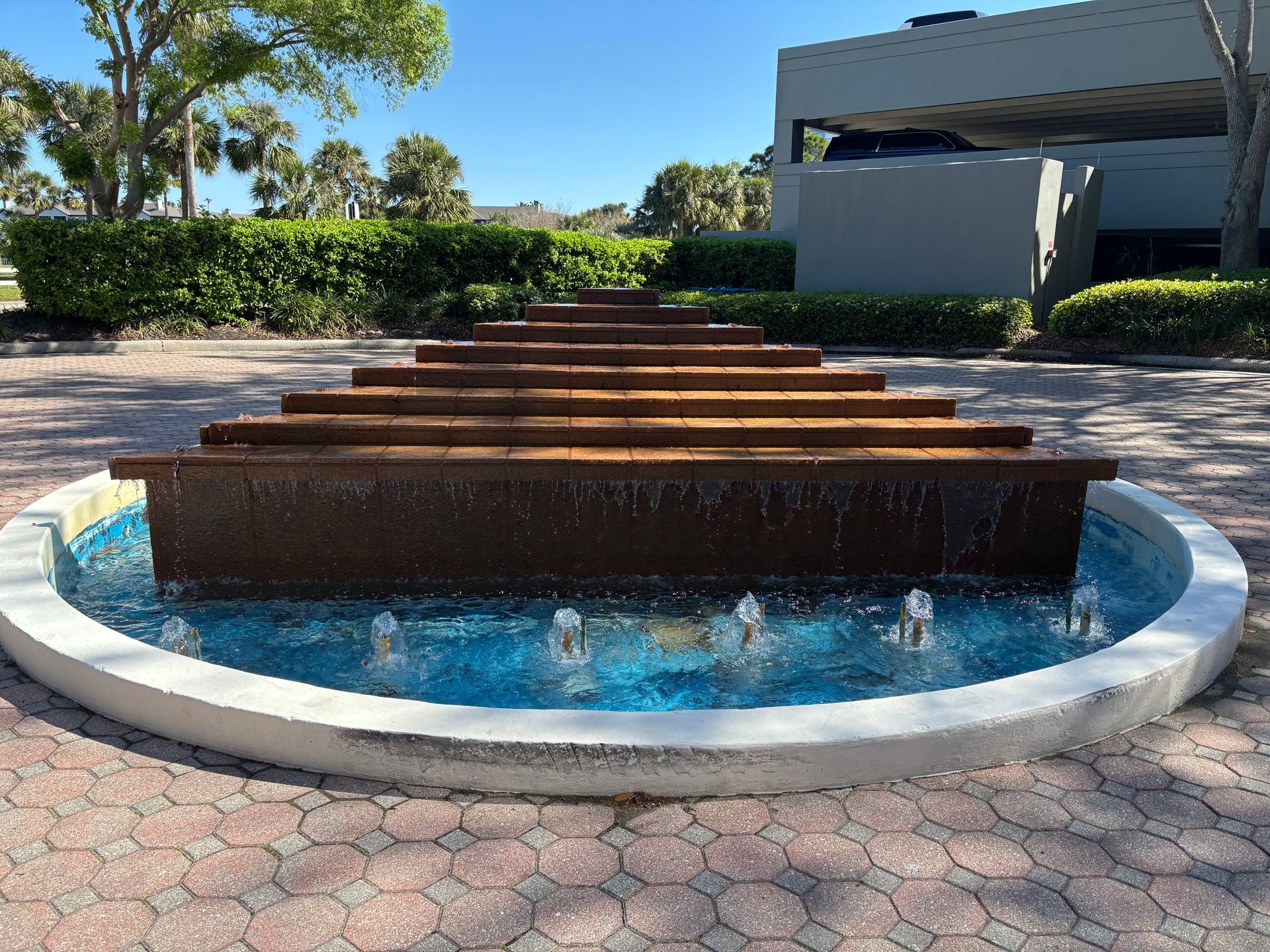 A tiered, rust-colored fountain with cascading water sits in a circular pool on a paved plaza in front of a building.
