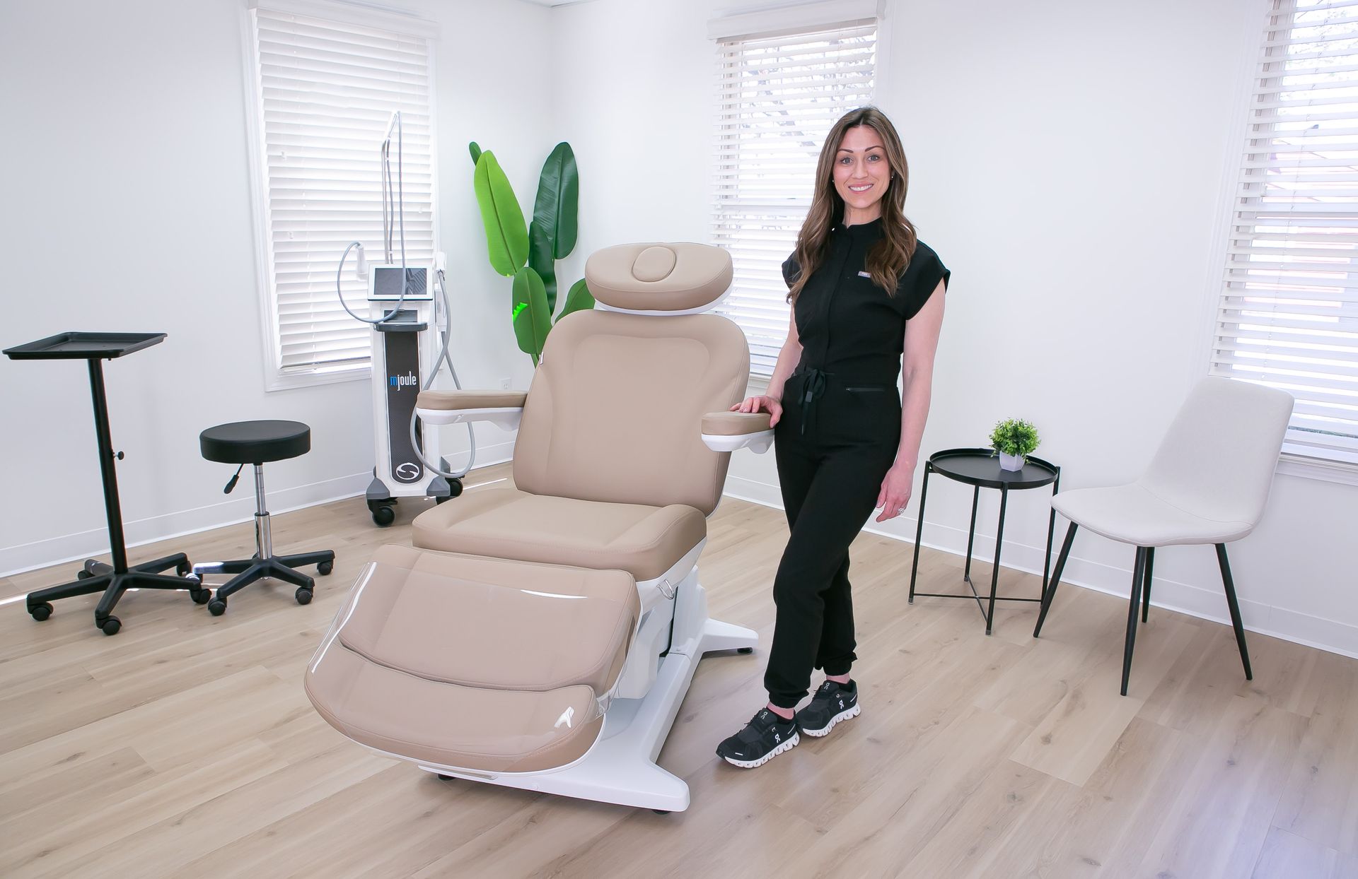 A woman in black scrubs stands beside a beige aesthetician chair in a treatment room.