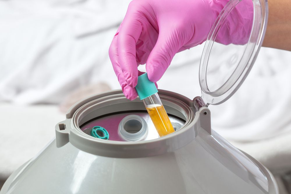 Gloved hand placing a test tube of yellow liquid into a centrifuge. Pink gloves, white background.