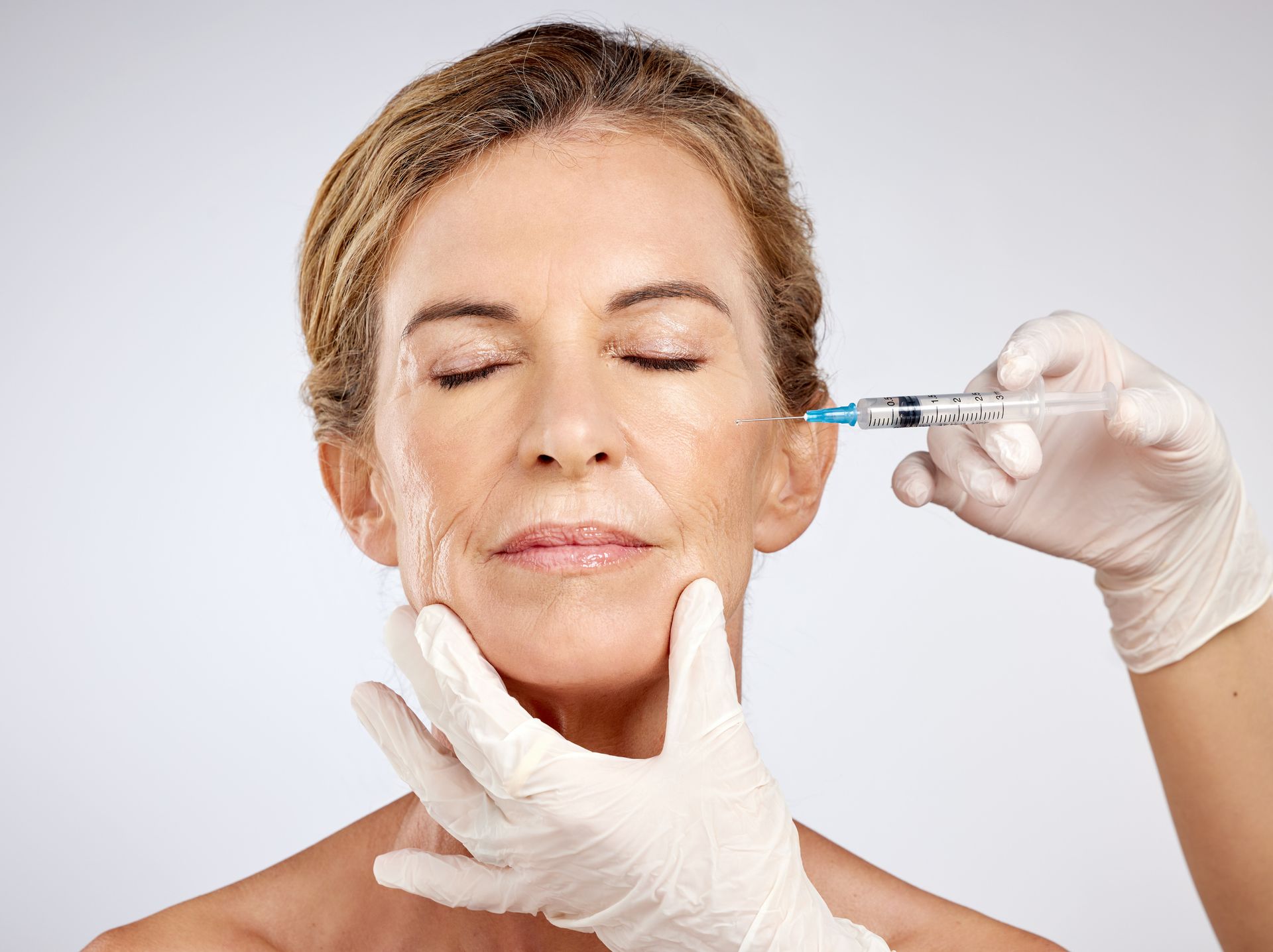 Woman receiving facial injections, doctor with black gloves, white room, green plant, beige chair.