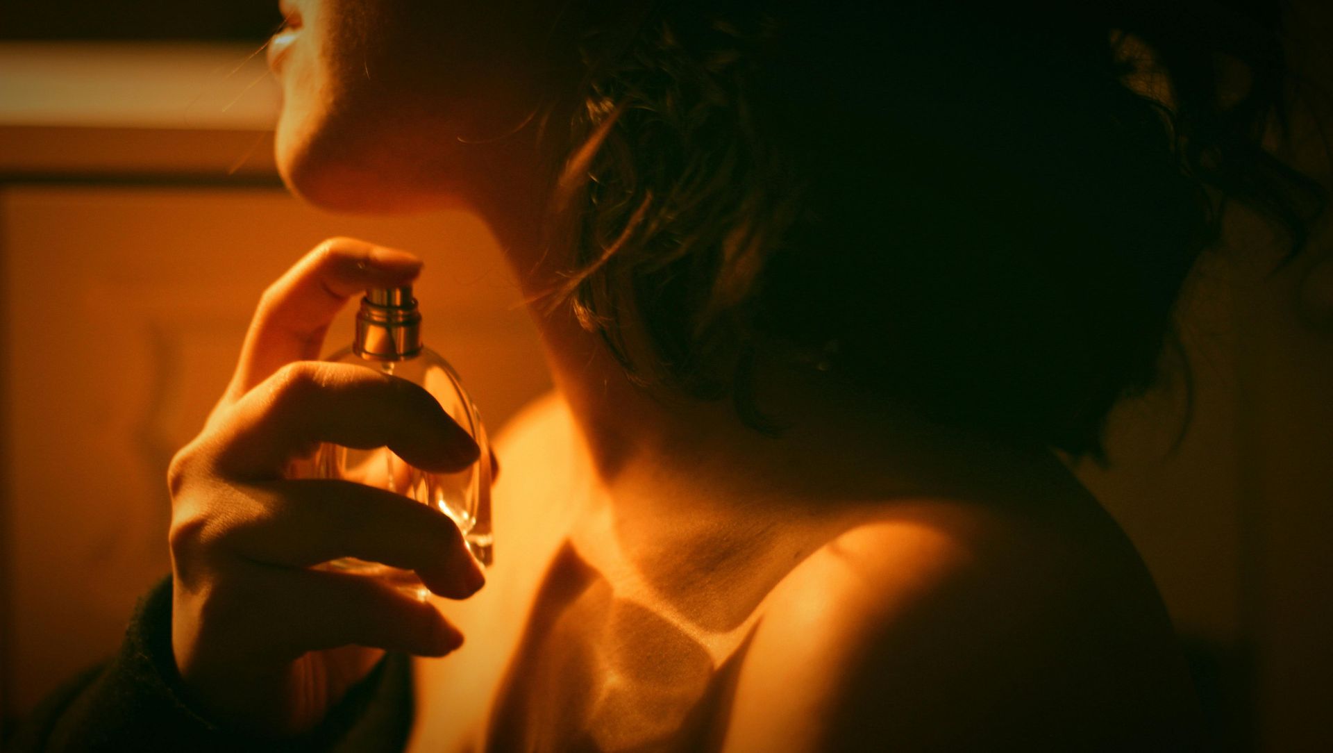 Woman spraying perfume on her neck. Warm lighting, hand holding a glass bottle.