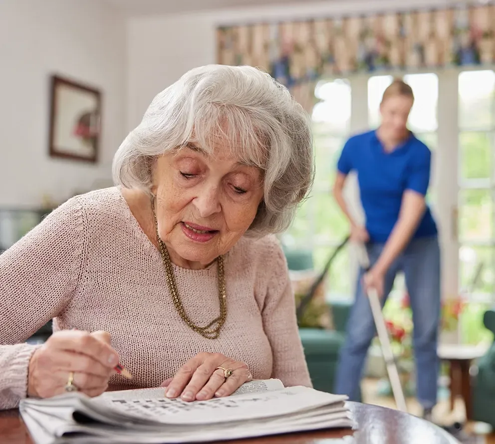 A Carer Vacuuming While A Lady Does a Crossword — Ani's Care Pty Ltd In Millner, NT