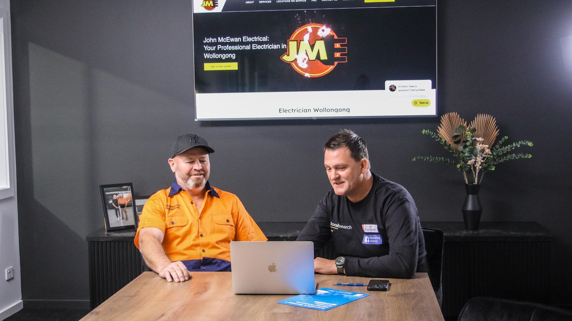 Two Men Seated At A Table, Looking At A Laptop. One Wears Work Attire, The Other A Black Shirt. A Screen Displays A Logo