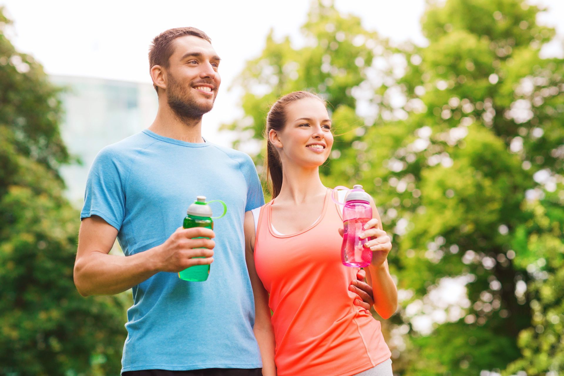 A man and woman smiling in a park, holding water bottles, and looking off-camera.