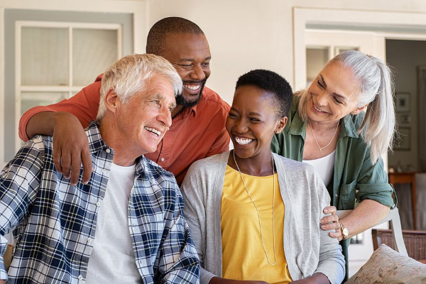 Four adults smiling, embracing. Two people have arms around each other. Inside a well-lit space.
