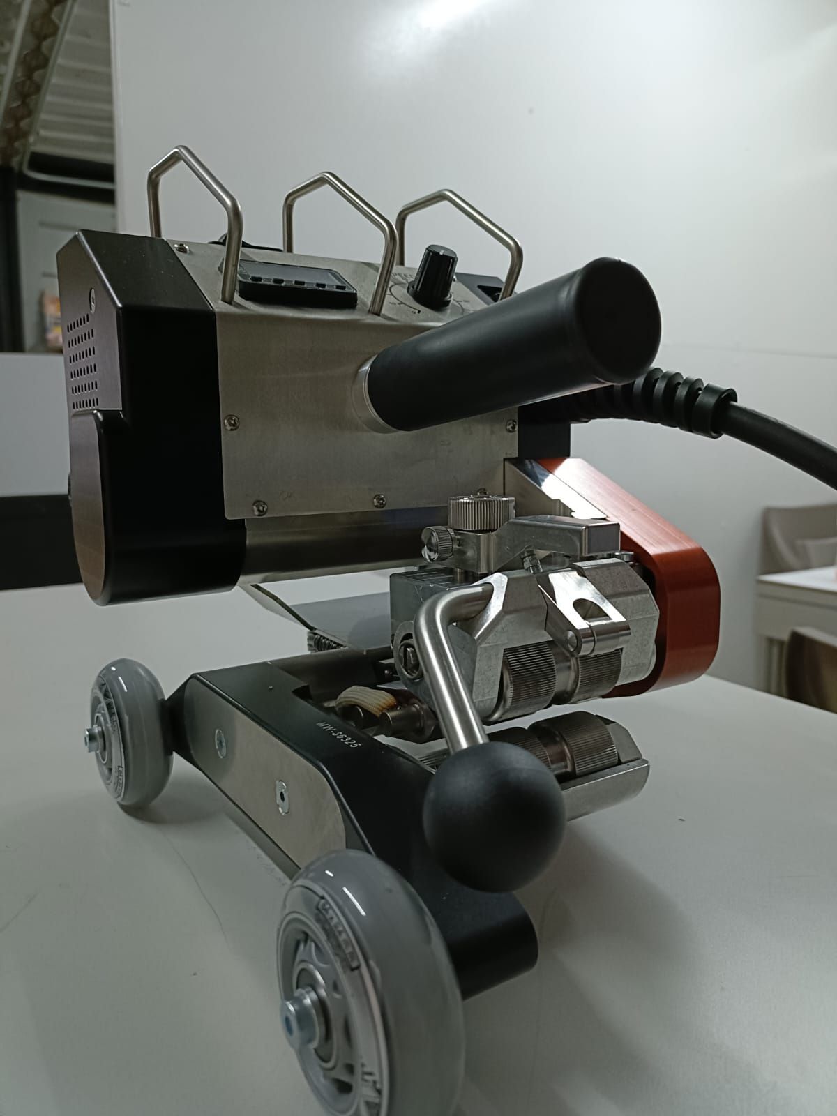 Máquina de soldar, de color gris y negro con una sección roja, sobre ruedas, asentada sobre una superficie blanca.