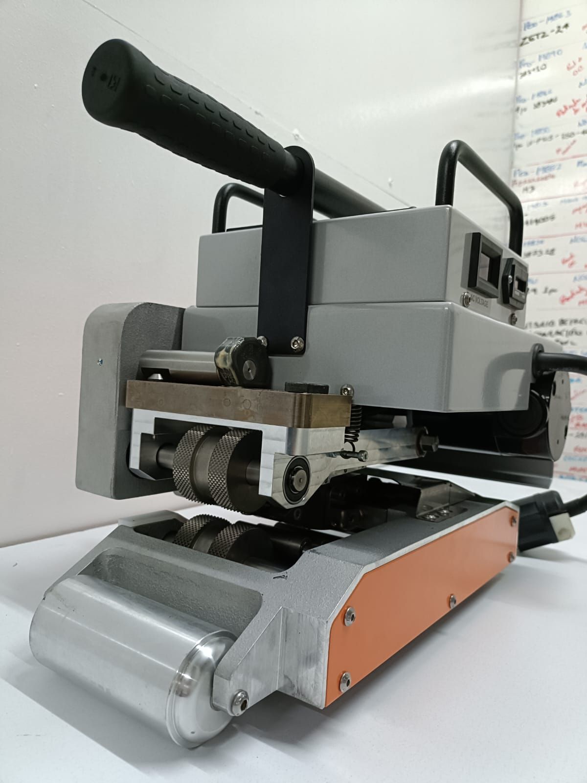 Soldadora de mano con mango negro y rodillos plateados. Cuerpo gris con detalles en naranja.