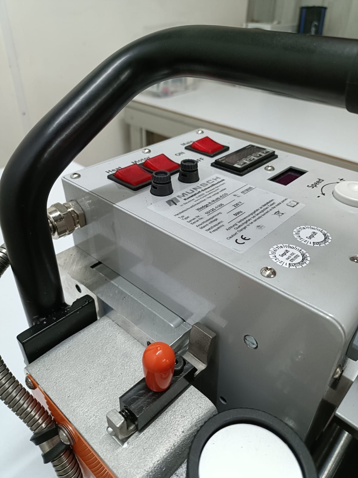 Primer plano de una máquina plateada con mango negro, interruptores rojos y una perilla naranja.
