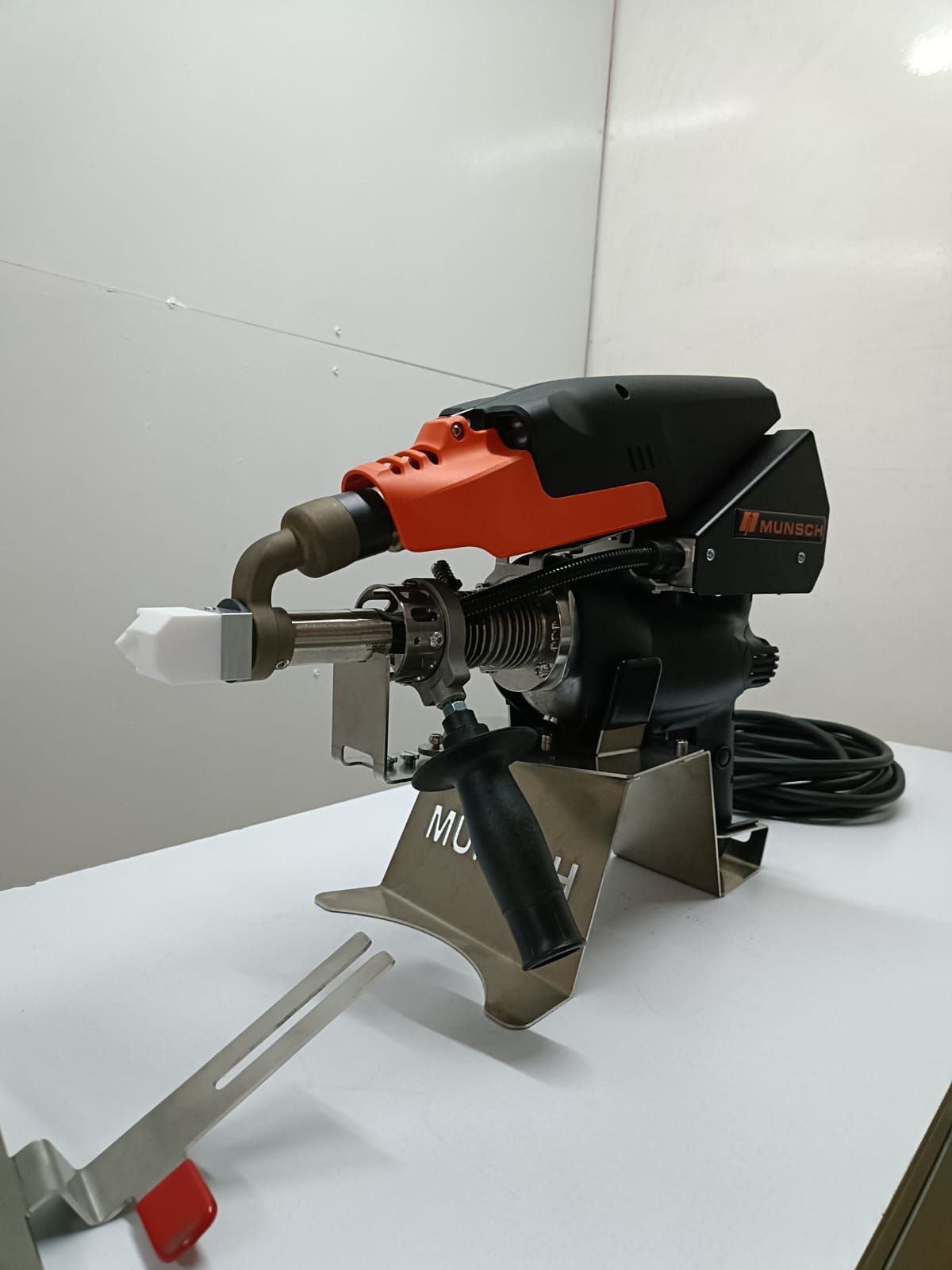 Máquina de soldar de plástico negro y naranja sobre soporte plateado, superficie blanca, en interiores.