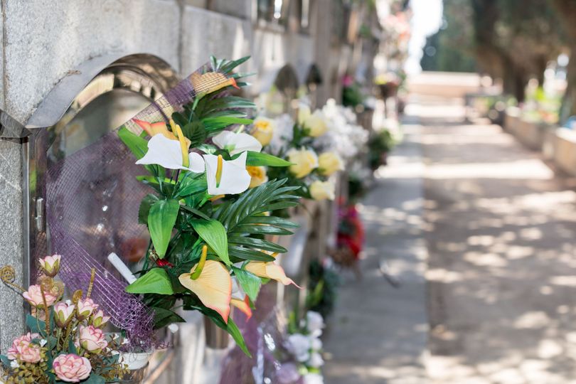 Una persona in abito nero tiene in mano una rosa bianca durante una cerimonia funebre con candele e composizioni floreali.