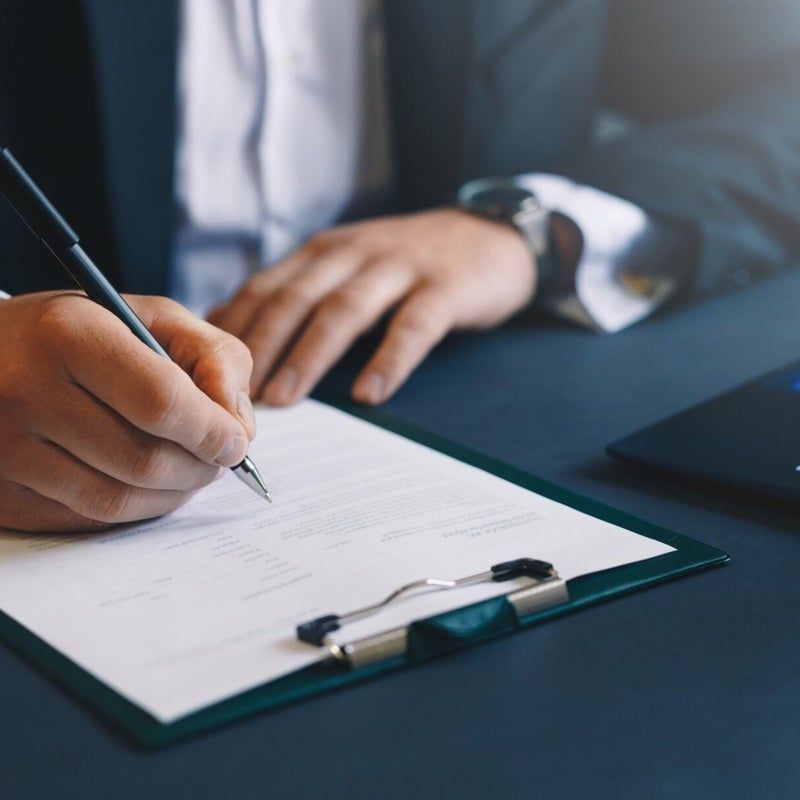 Persona in giacca e cravatta che scrive su un documento tenuto in una cartellina, su una scrivania blu scuro.