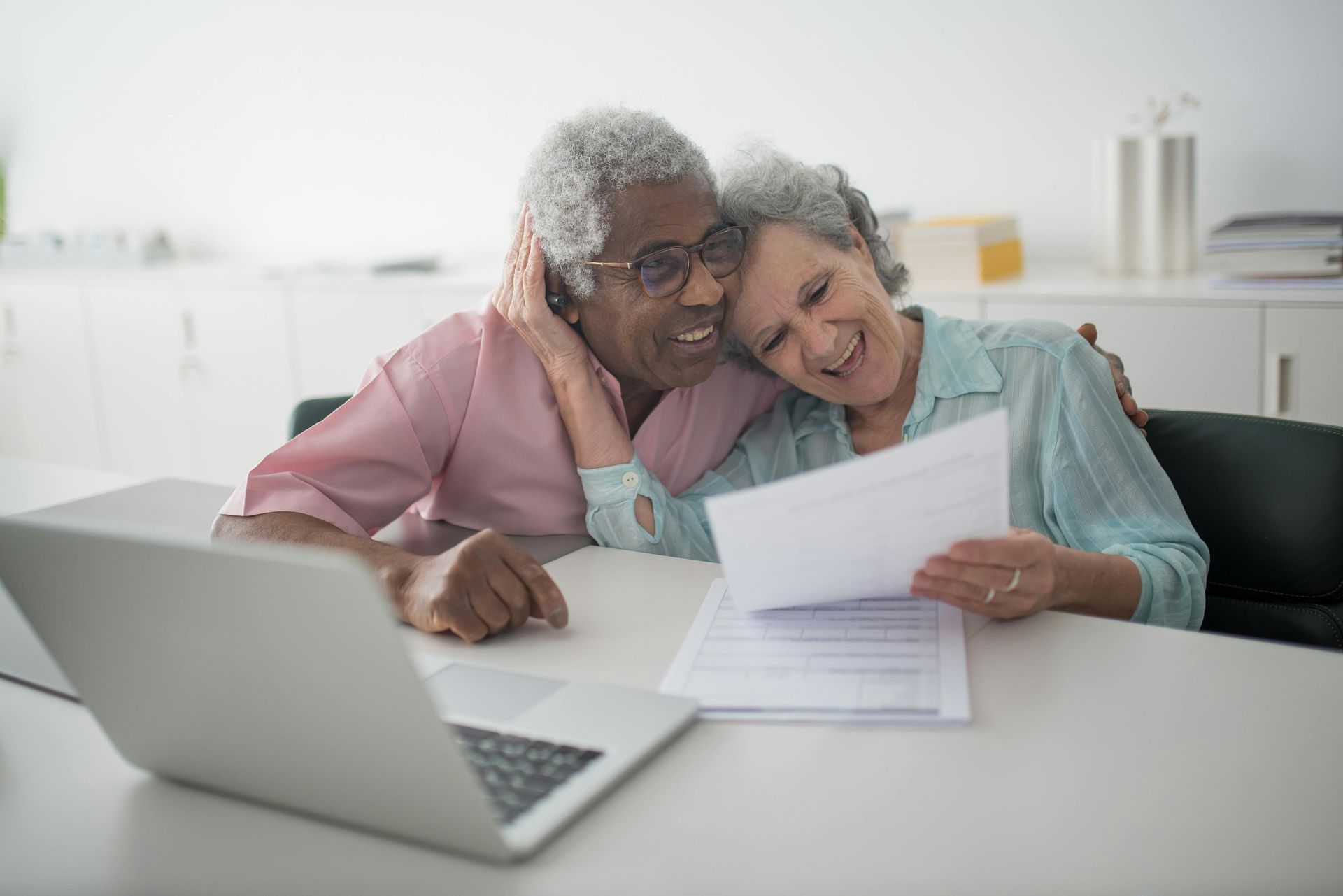 Smiling elderly couple, embracing, reading documents together at a table with a laptop.