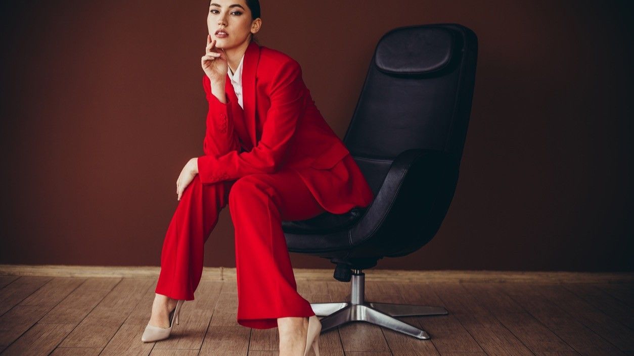 Woman in red suit sits in black office chair. Leaning against a brown wall, she looks at the camera.
