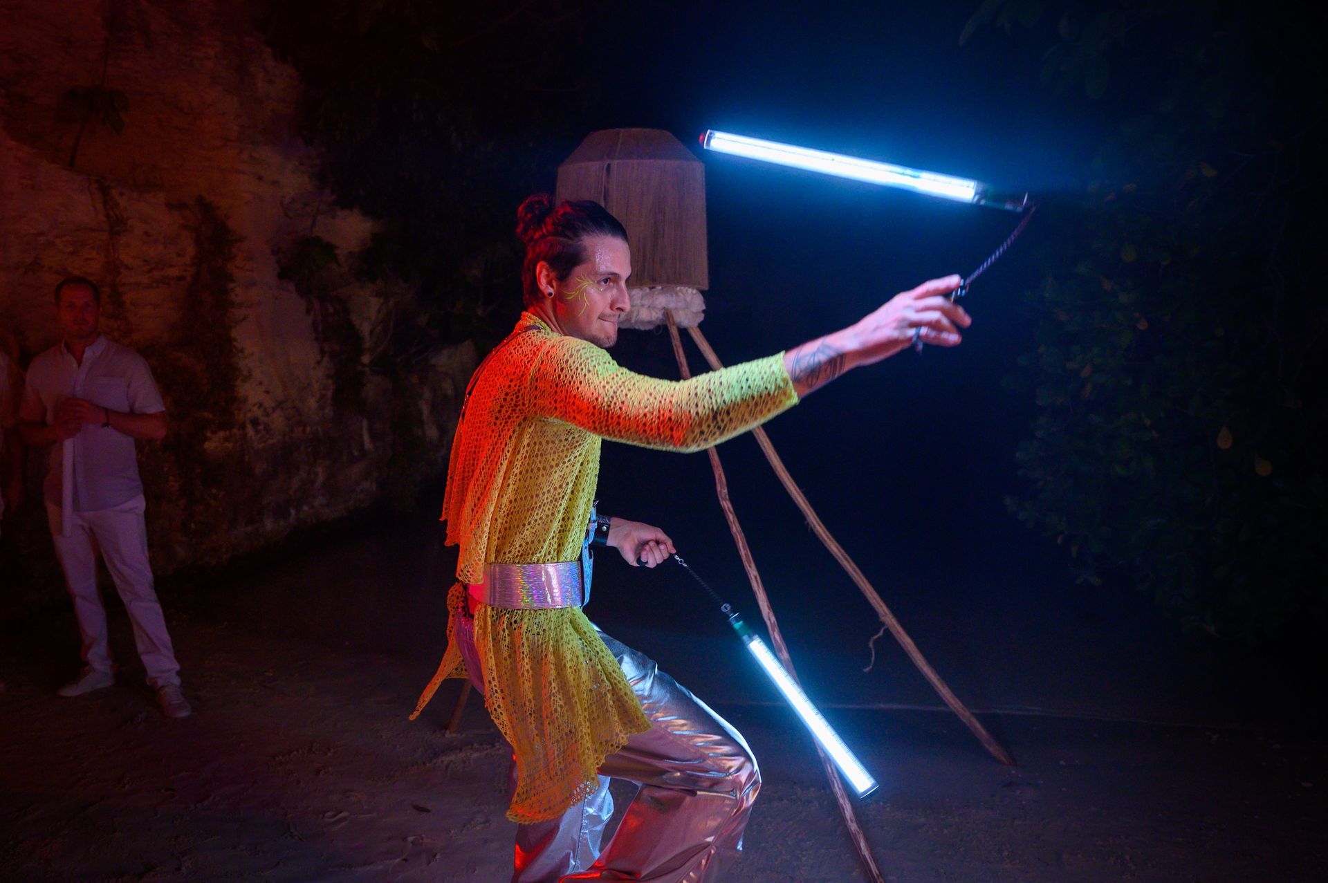A man in a yellow dress is holding two lightsabers in a dark room.