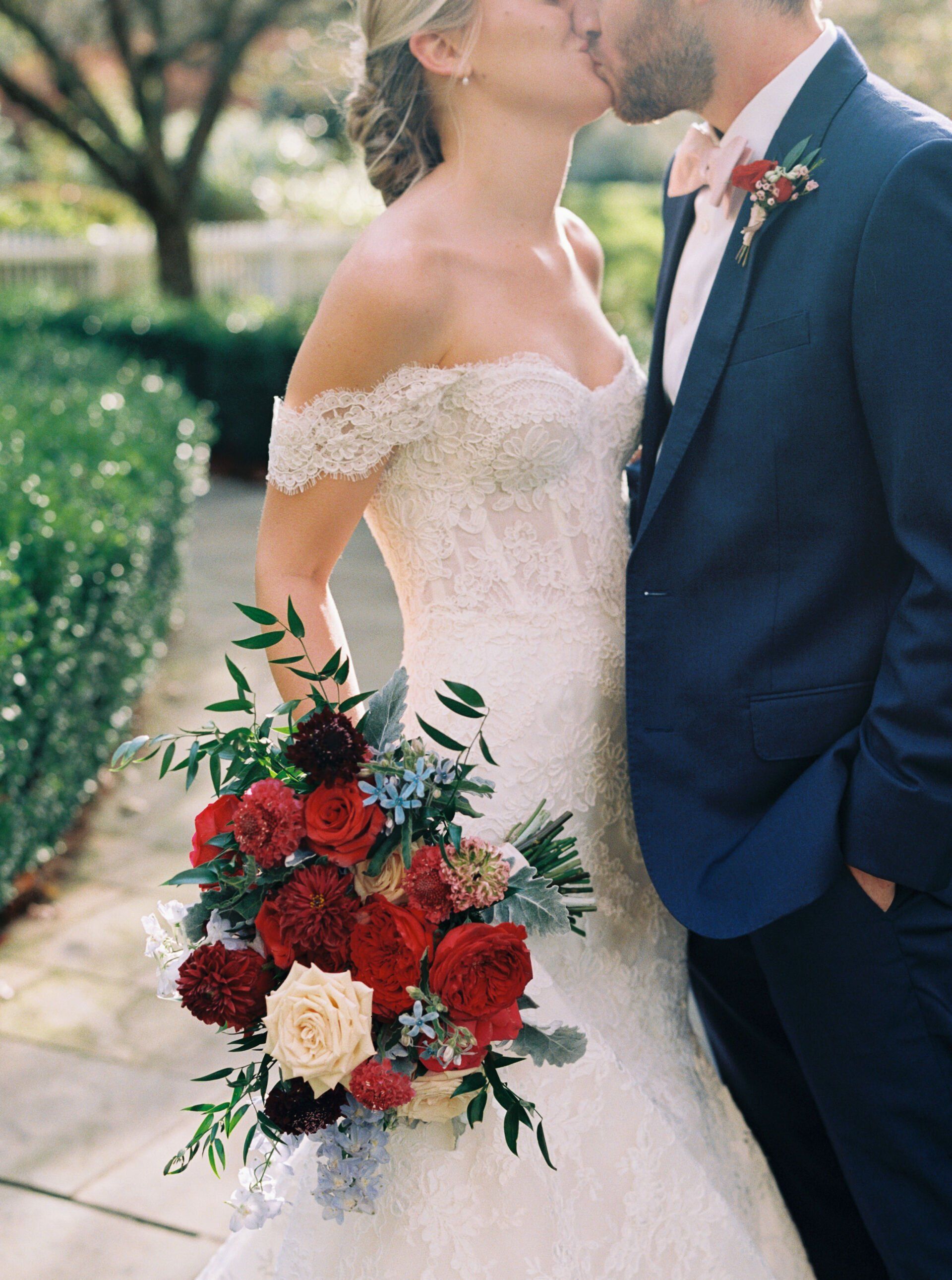 A bride and groom kissing while the bride is holding a bouquet of red flowers.