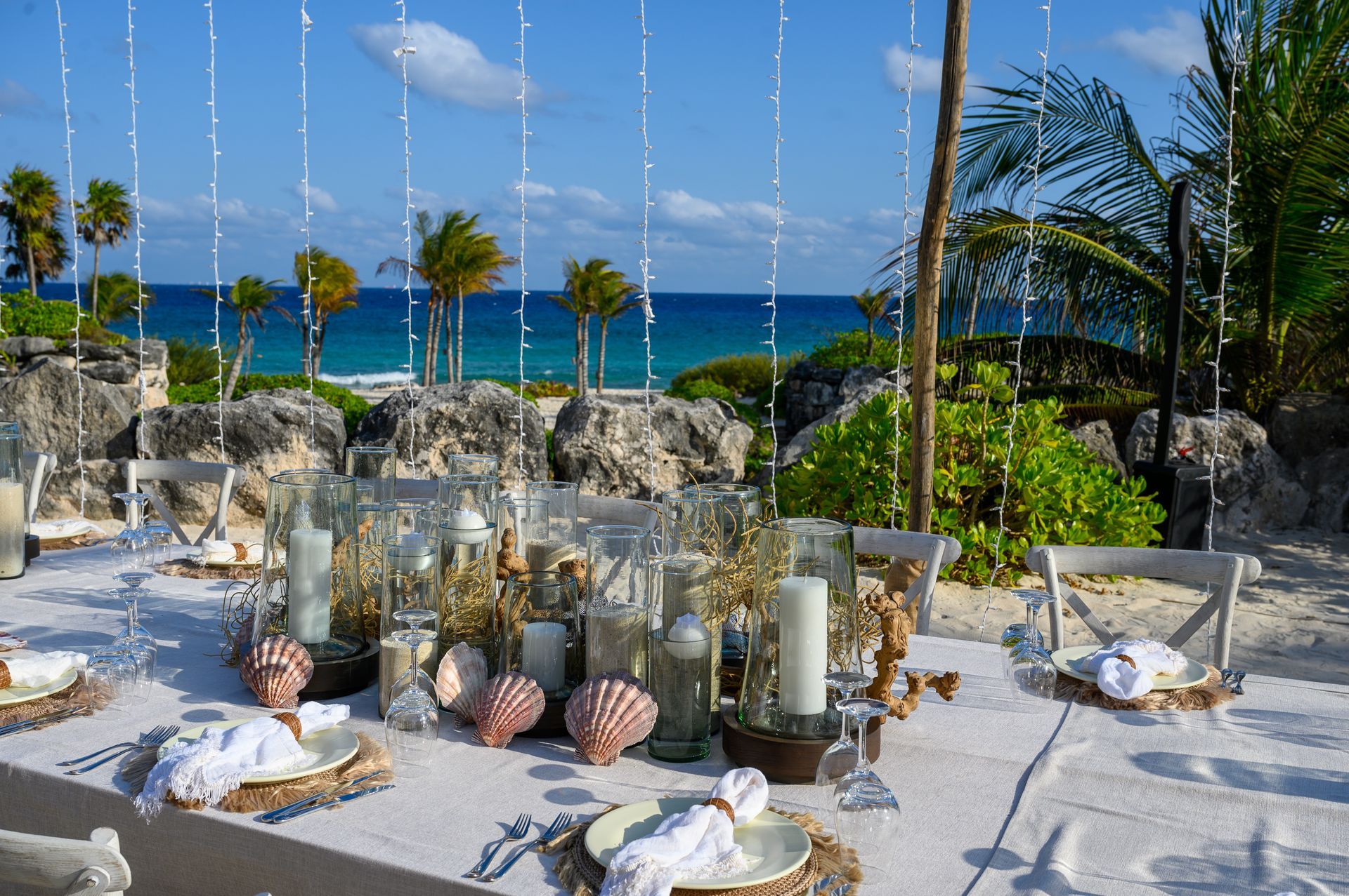 A long table with plates , candles , and seashells on it in front of the ocean.