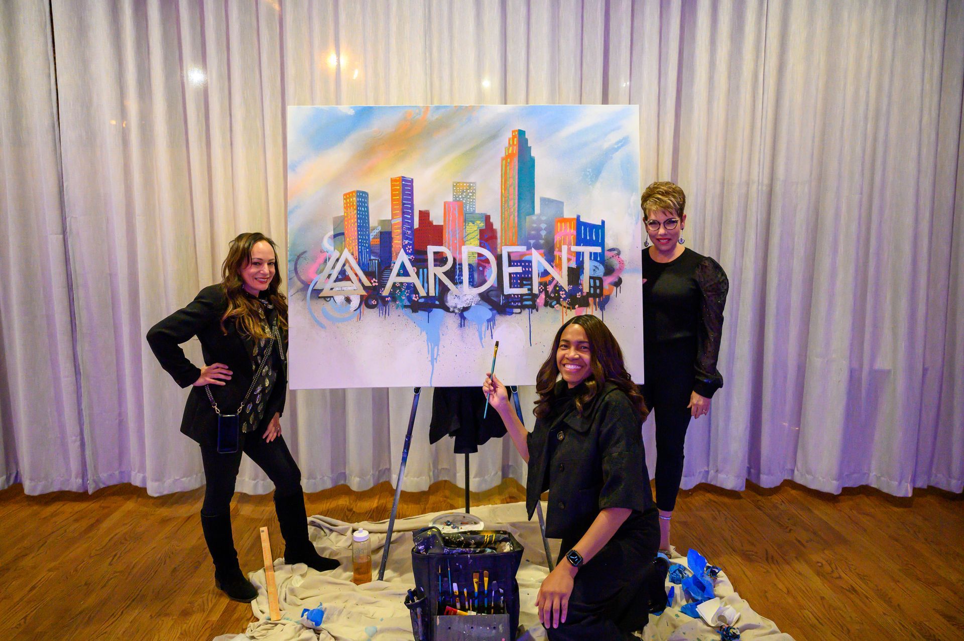 Three women are standing around a large painting of a city skyline.