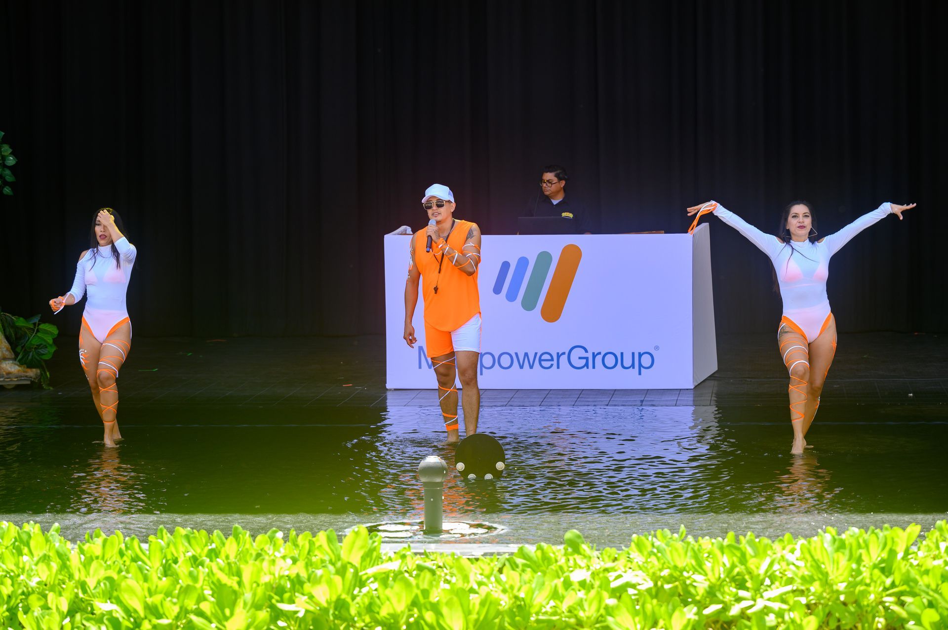 A man is singing into a microphone while two women are dancing in the water.
