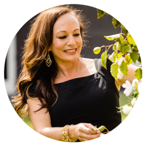 A woman in a black dress is standing next to a tree and smiling.