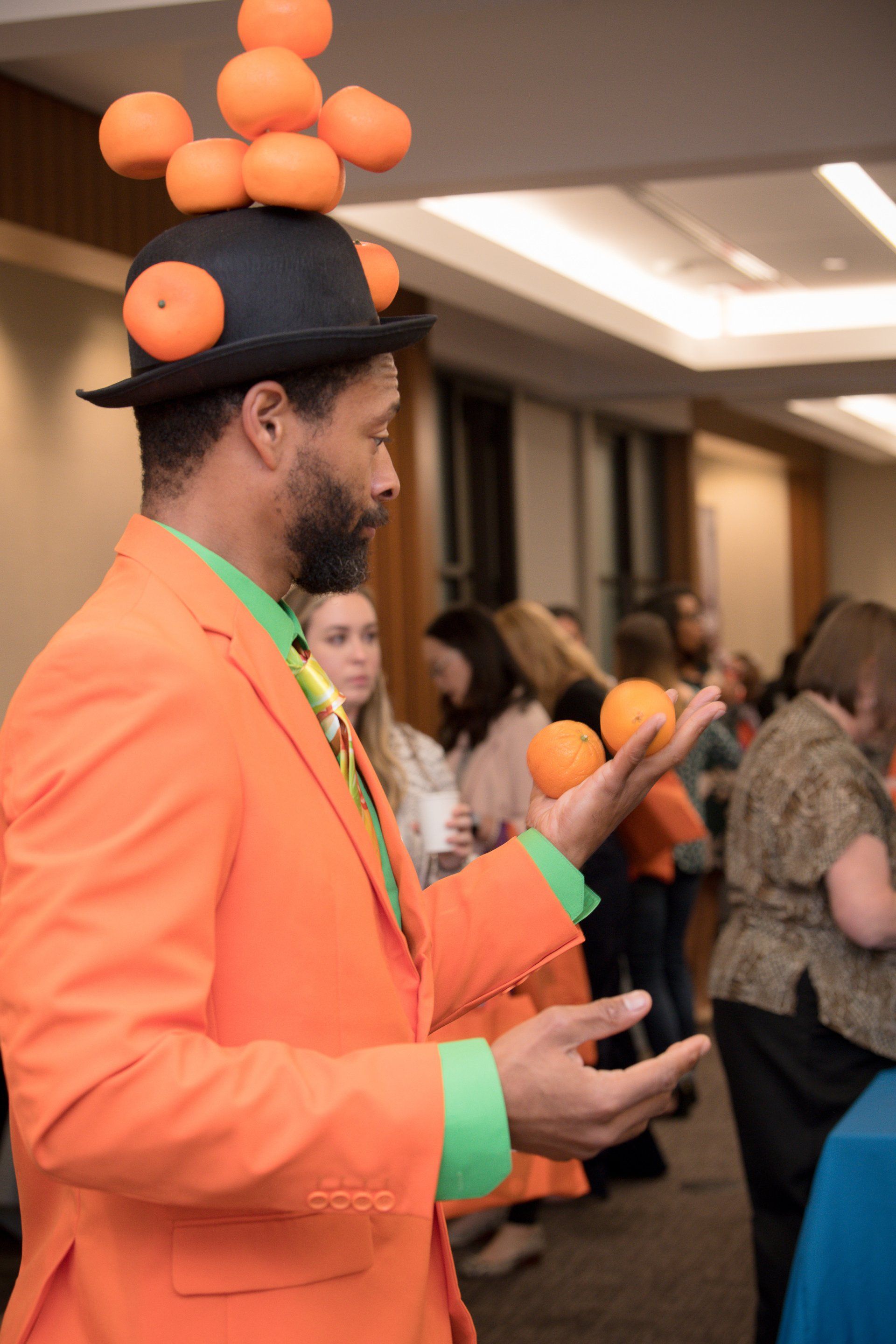 A man in an orange suit is juggling oranges on his hat.