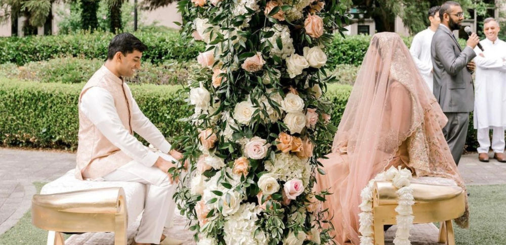 A bride and groom are sitting on a bench in front of a flower arrangement.