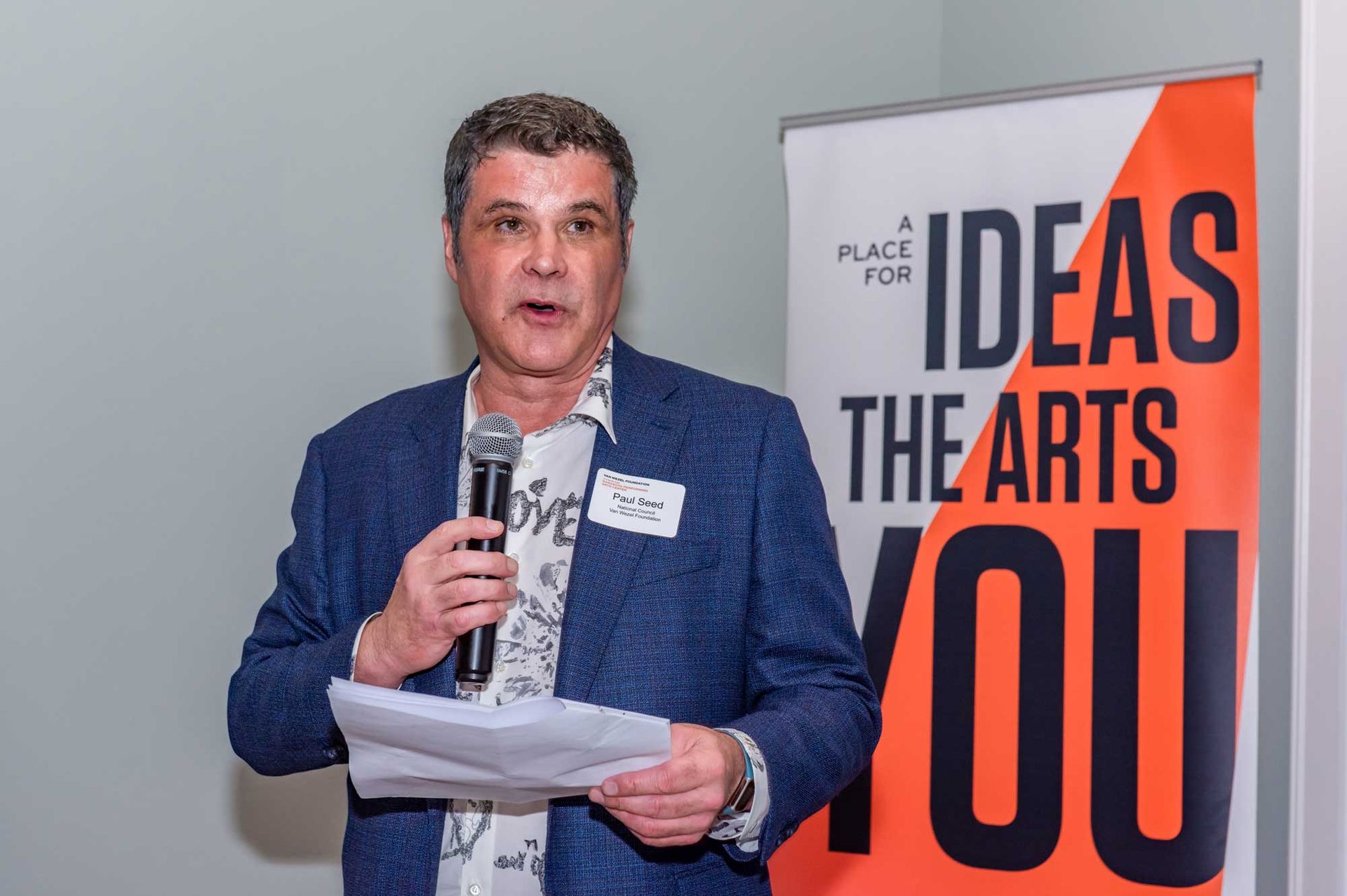 Man in blazer speaks into microphone at an arts event with banner 