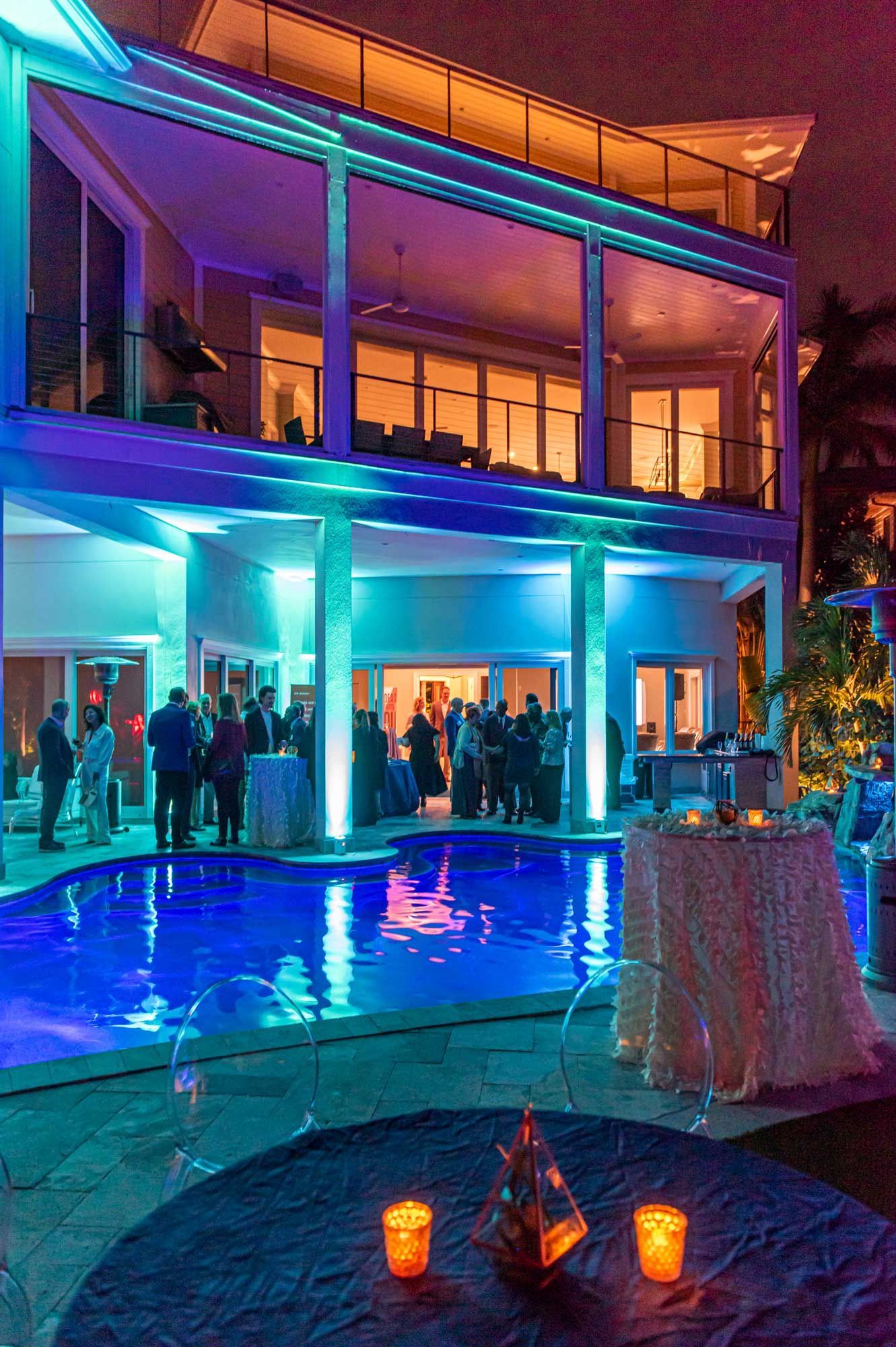 A group of people are standing around a swimming pool in front of a large house.