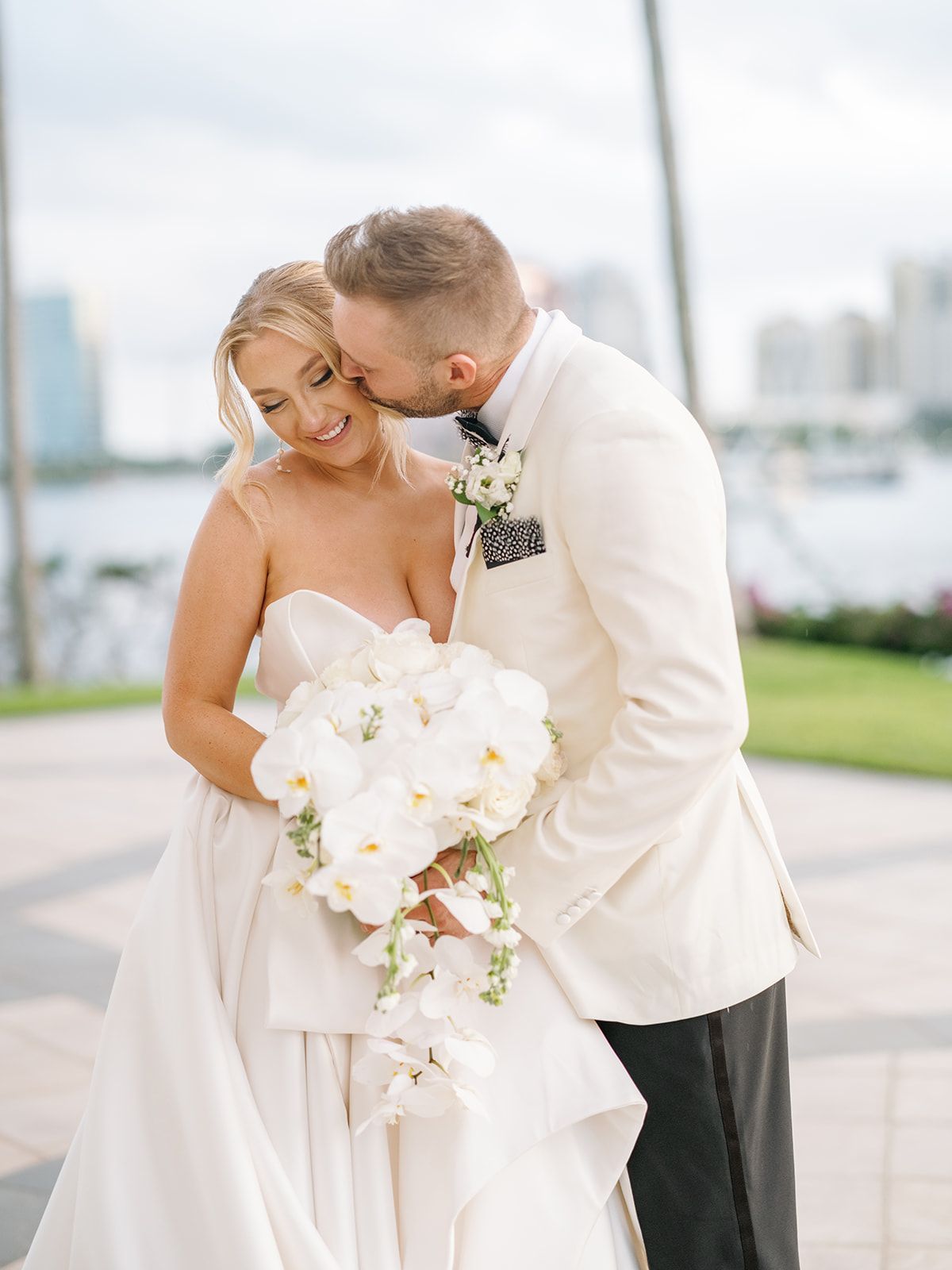 A bride and groom are posing for a picture and the groom is kissing the bride on the forehead.