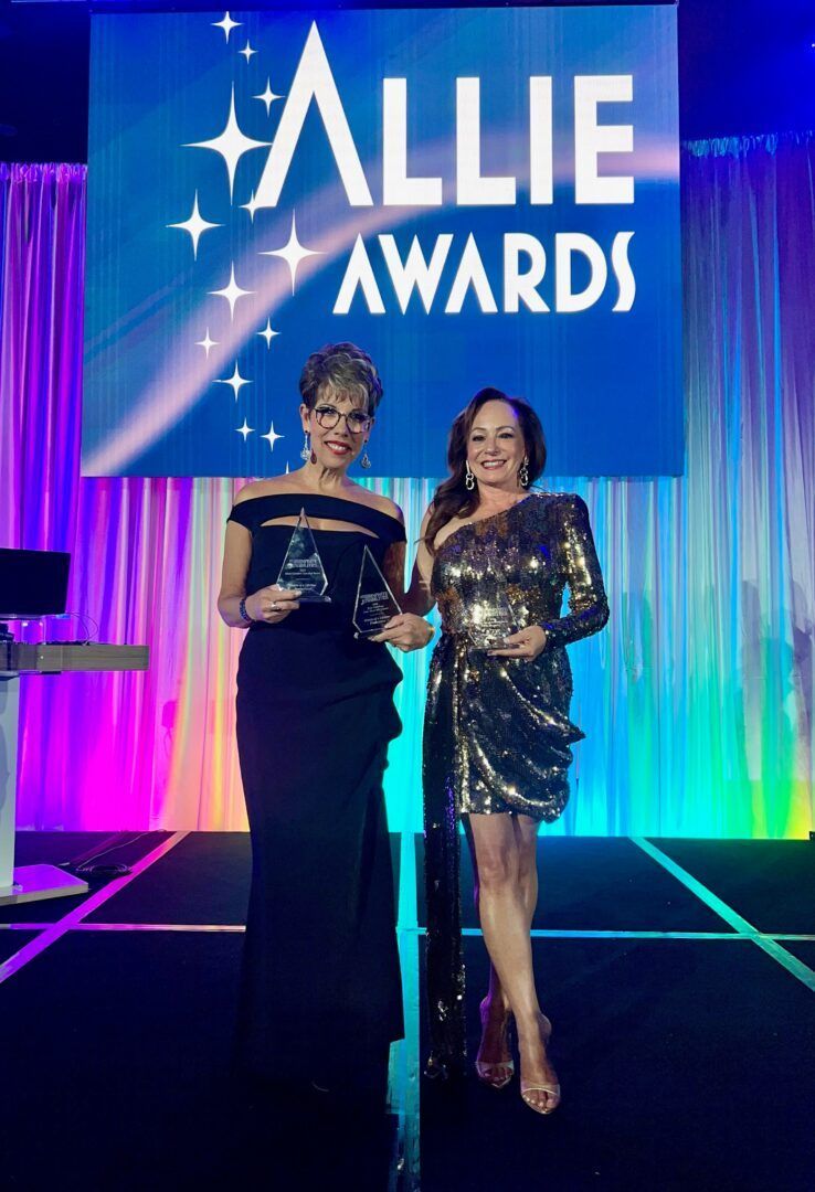 Two women holding awards at the Allie Awards. One wears a dark gown, the other a gold dress. Stage background.