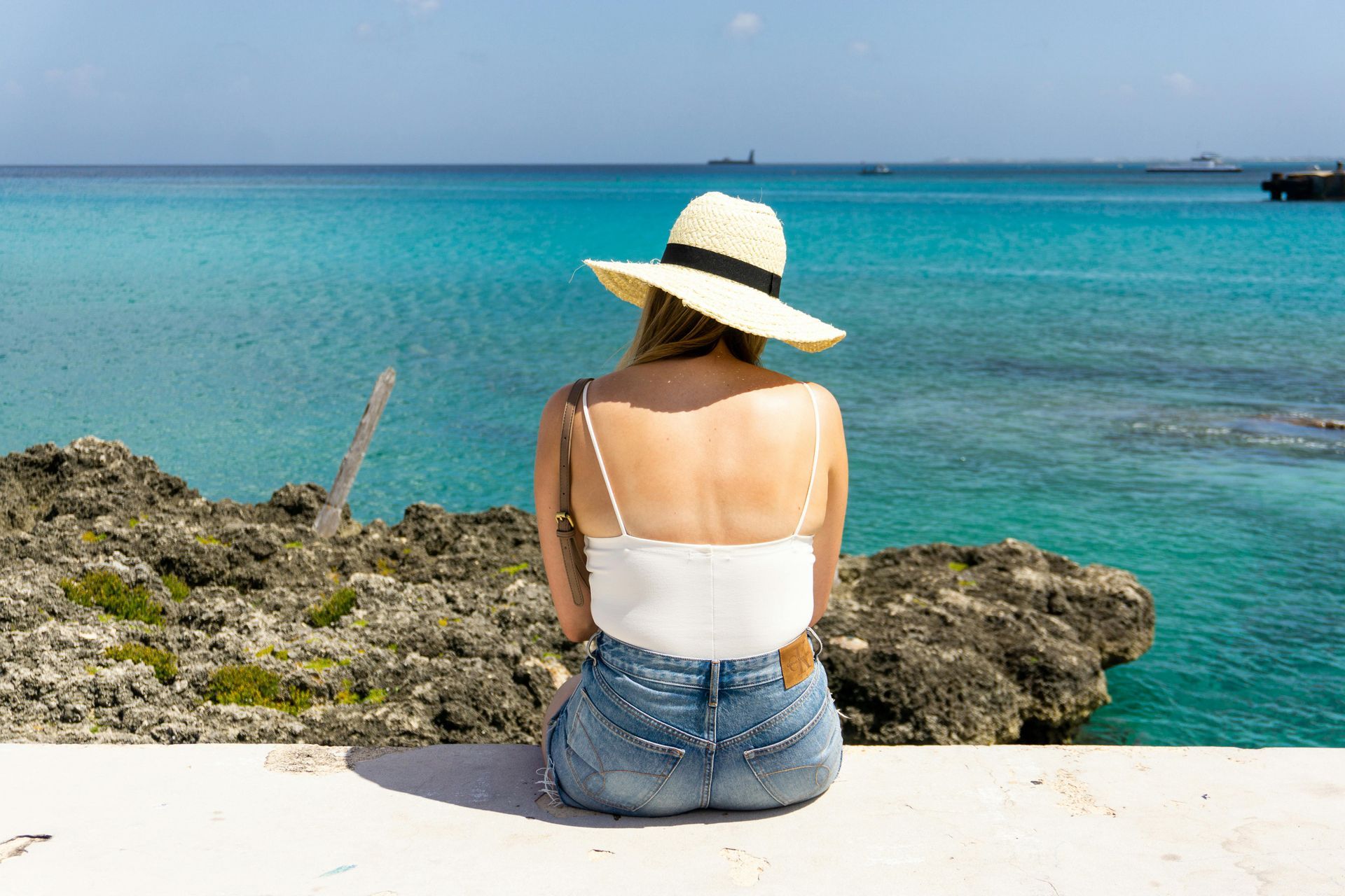 Woman in hat sits on a wall overlooking turquoise ocean.