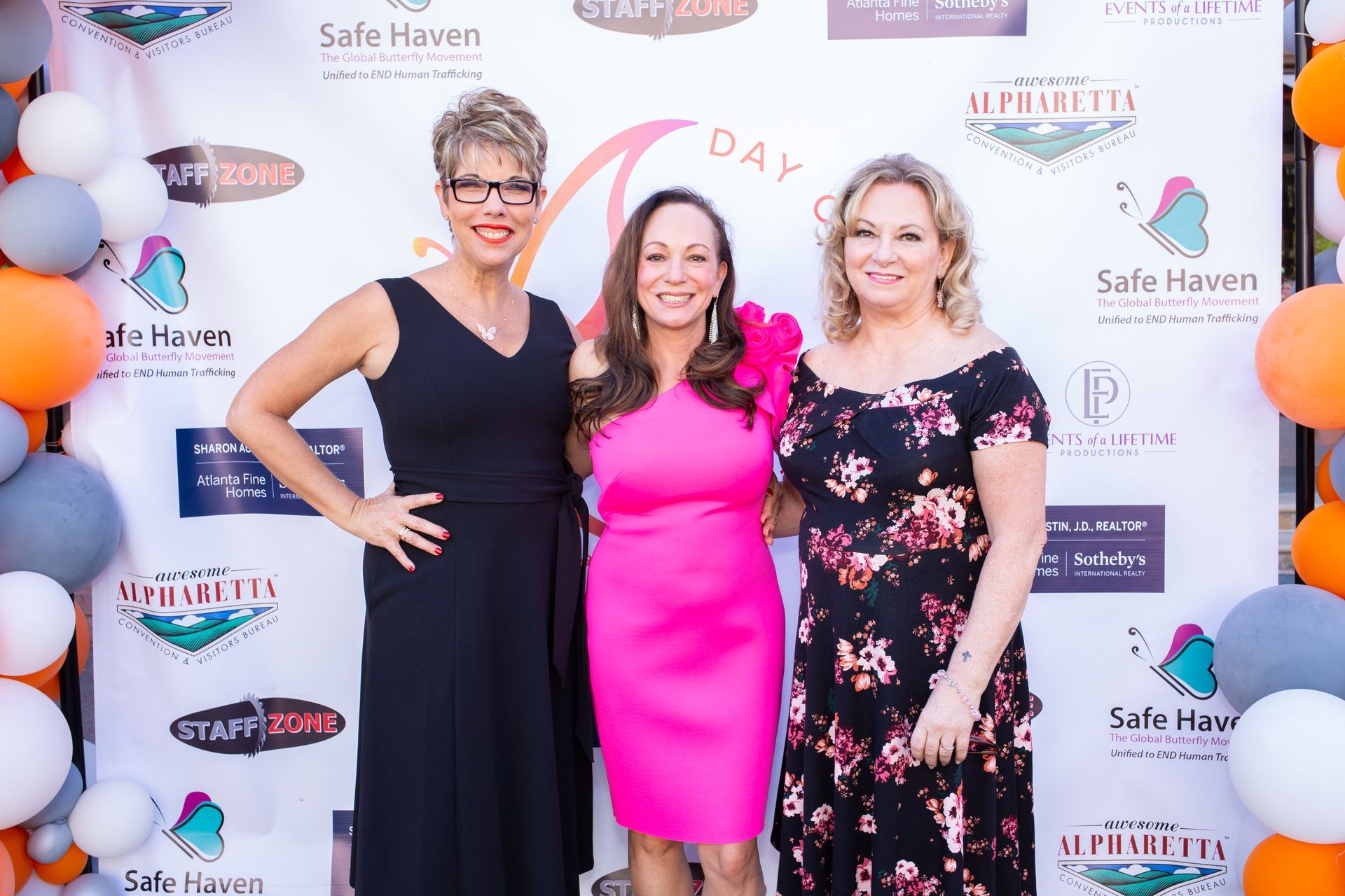 Three women are posing for a picture in front of a backdrop with balloons.