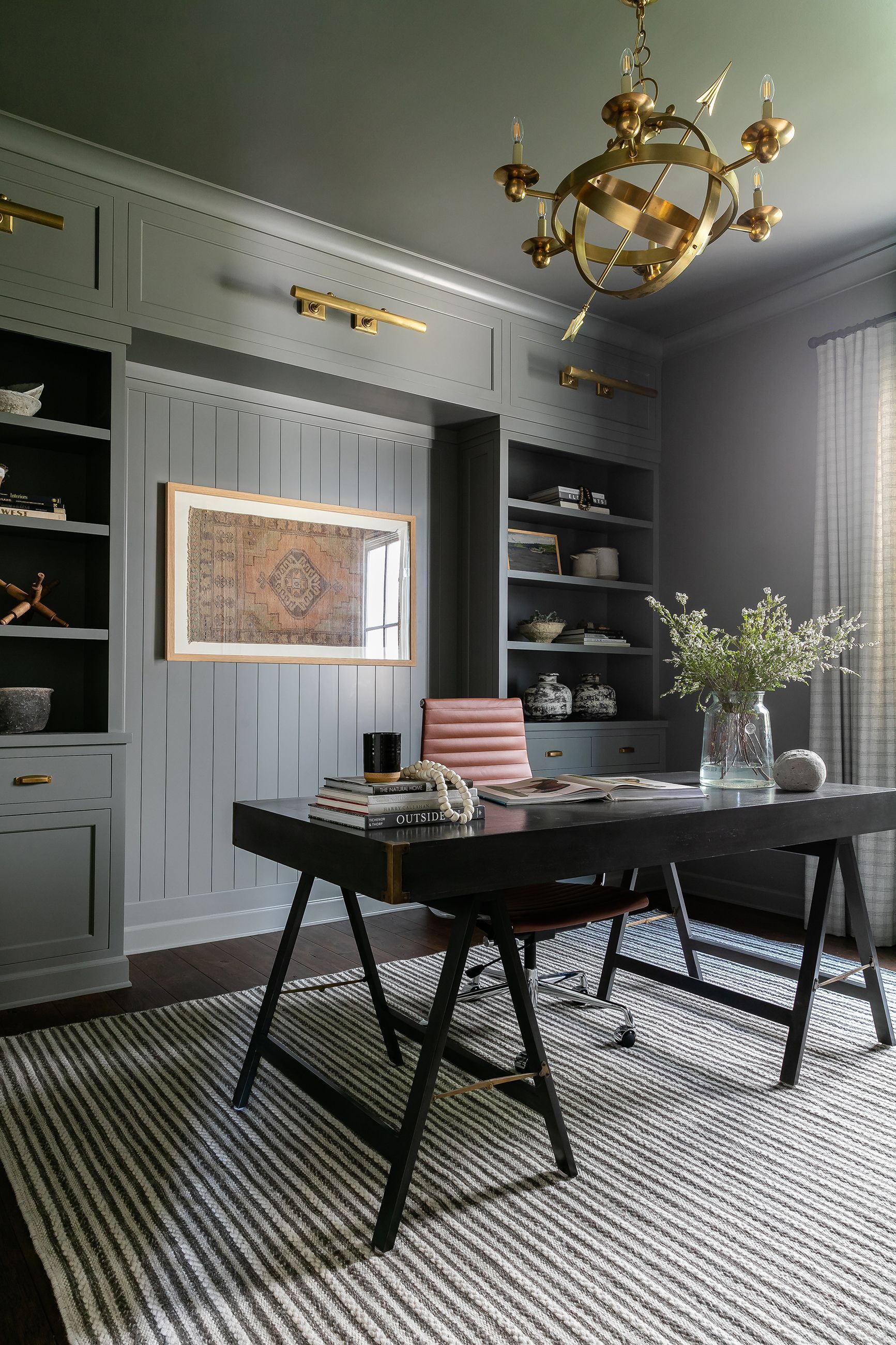 Gray home office with dark desk, shelves, patterned rug, and gold chandelier.