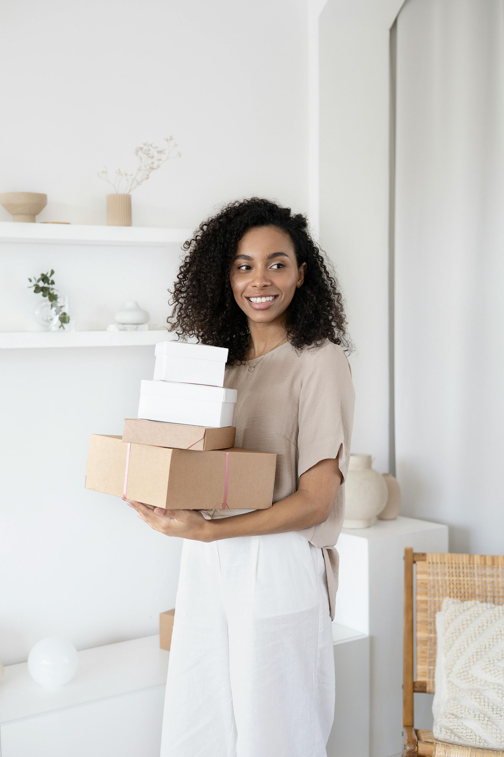 Woman holding a stack of boxes, smiling. Standing in a white room with a shelf, and decor.