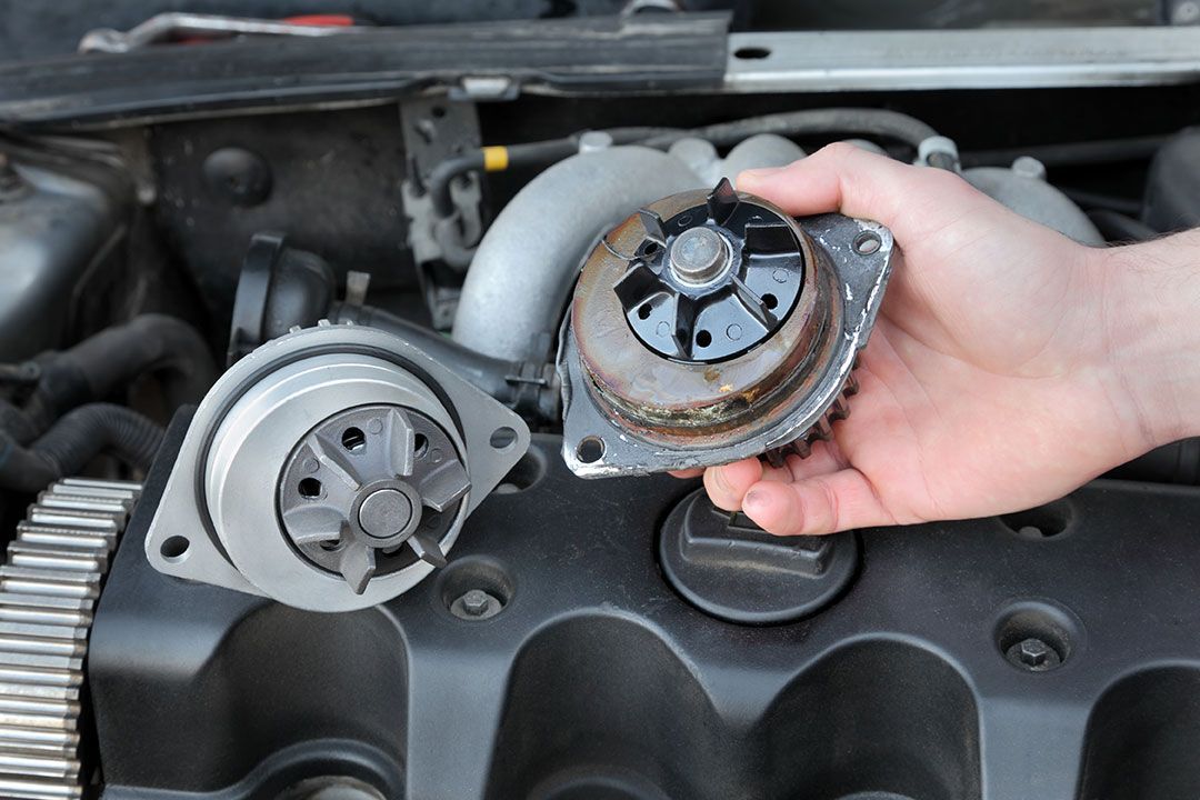 Hand holding a worn metal car engine part beside another mounted part in an open engine bay