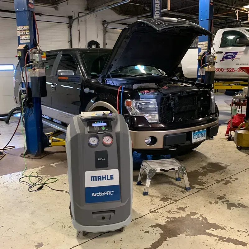 Pickup truck in a mechanic’s garage with hood open, next to a MAHLE air-conditioning service machine.