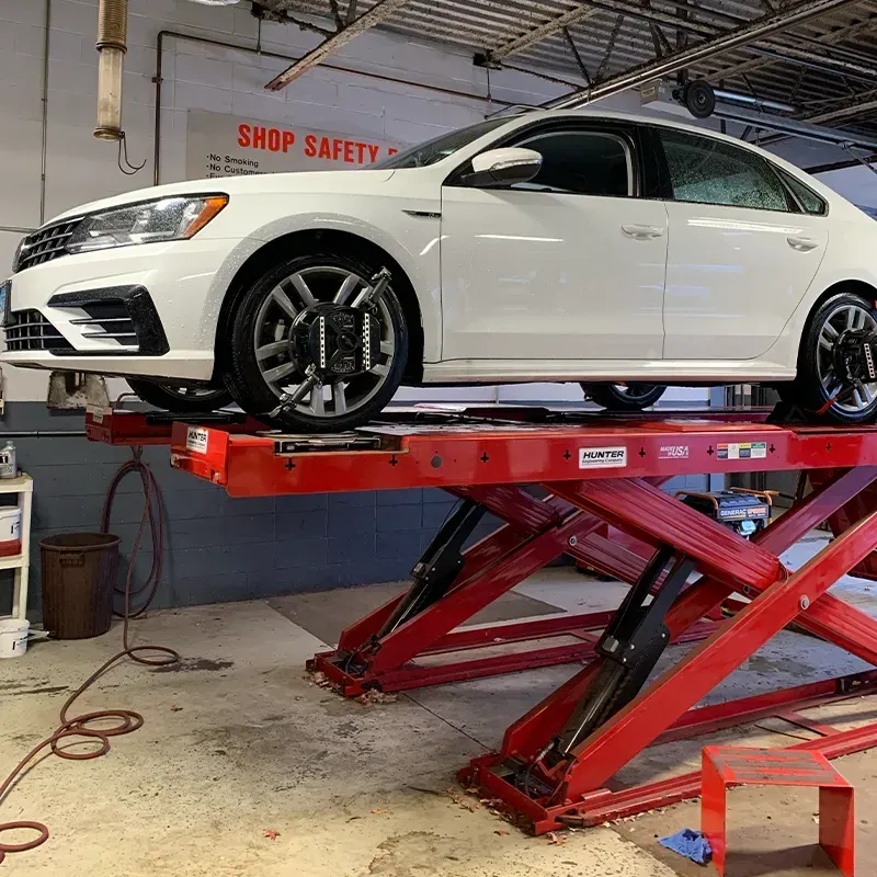 White sedan on a red scissor lift in an auto repair shop with a “Shop Safety” sign on the wall