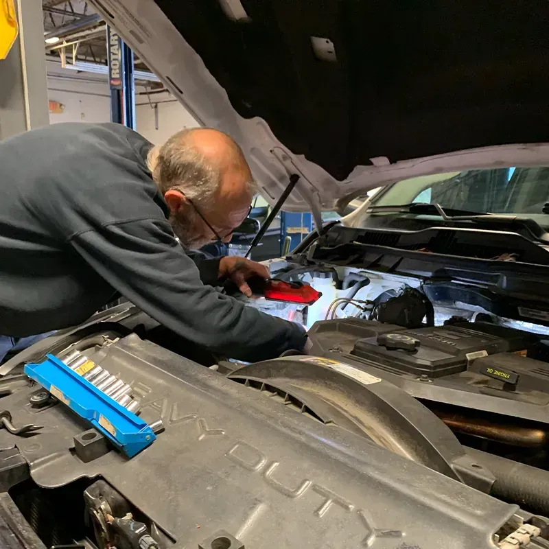 Mechanic working under a car hood in a garage, using a red tool near the engine.