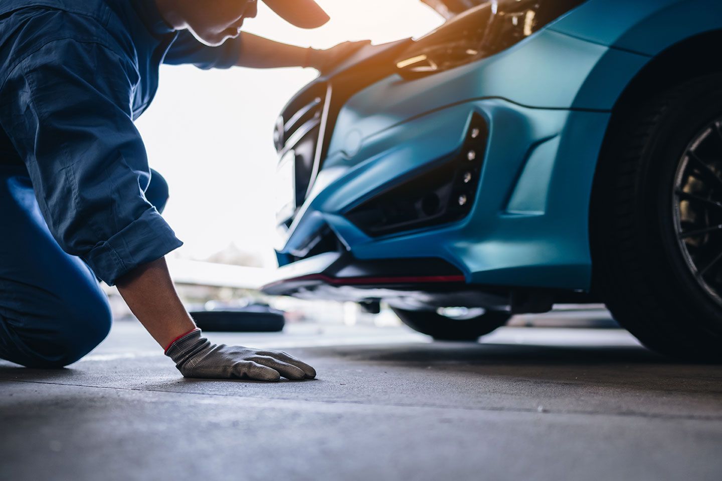 Mechanic kneeling beside a blue sports car, inspecting the front bumper in a garage